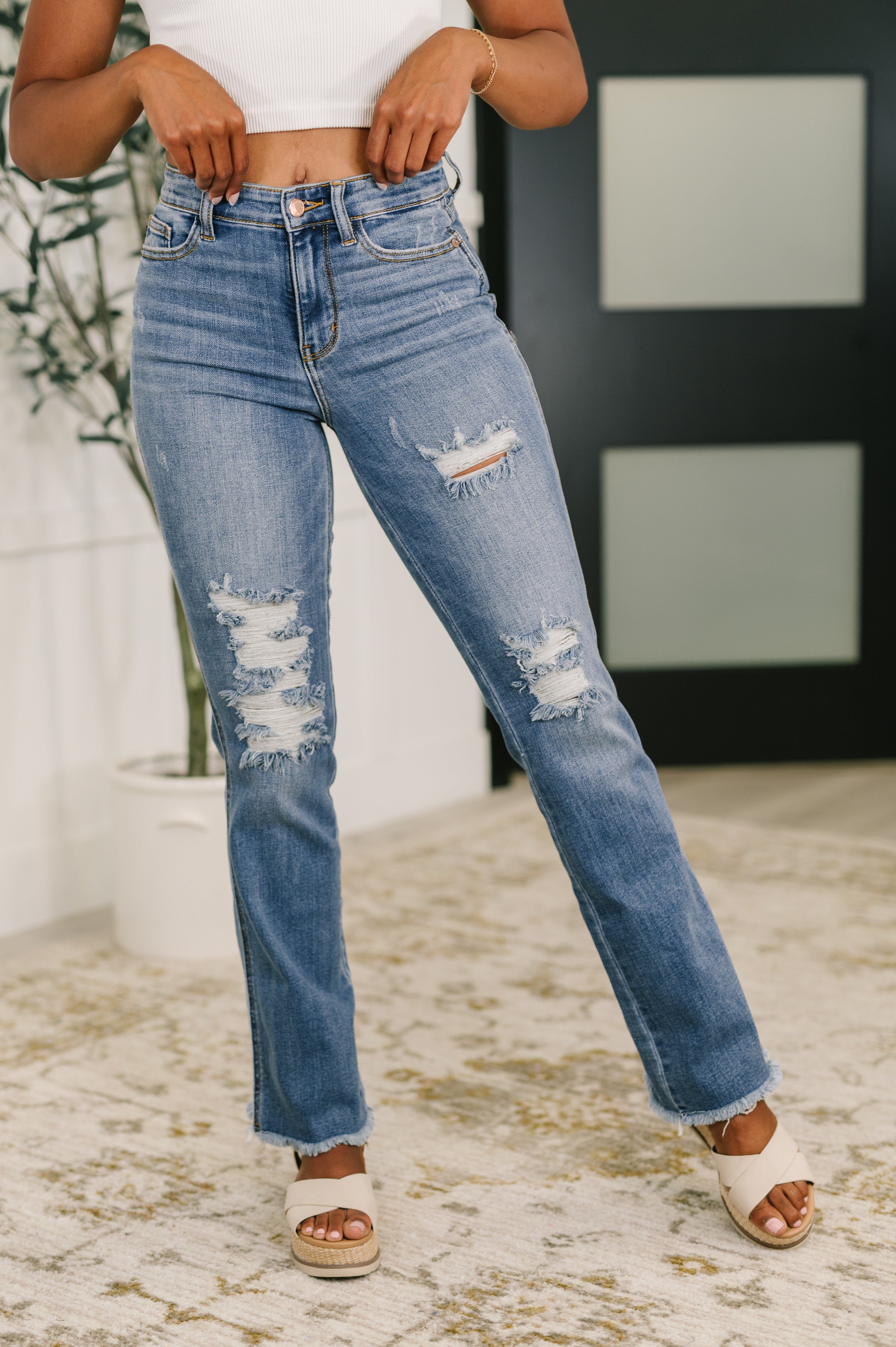 Front view of blue high-rise distressed bootcut jeans with ripped details and frayed hems worn with white sandals