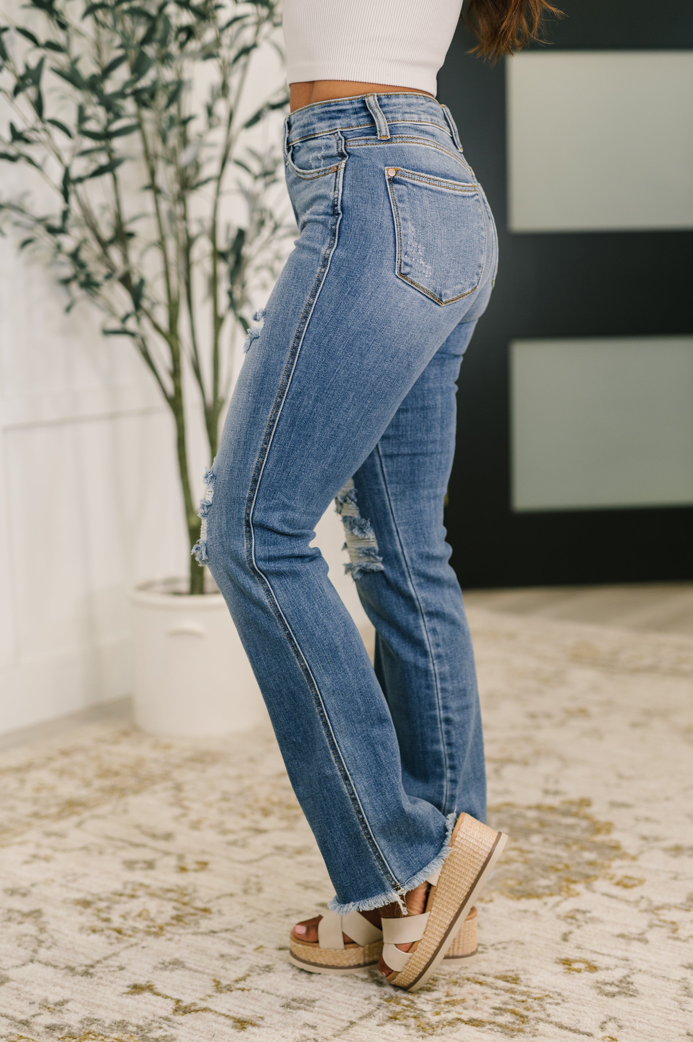 Side view of blue high-rise bootcut jeans with distressed knee details and frayed hems paired with white platform sandals
