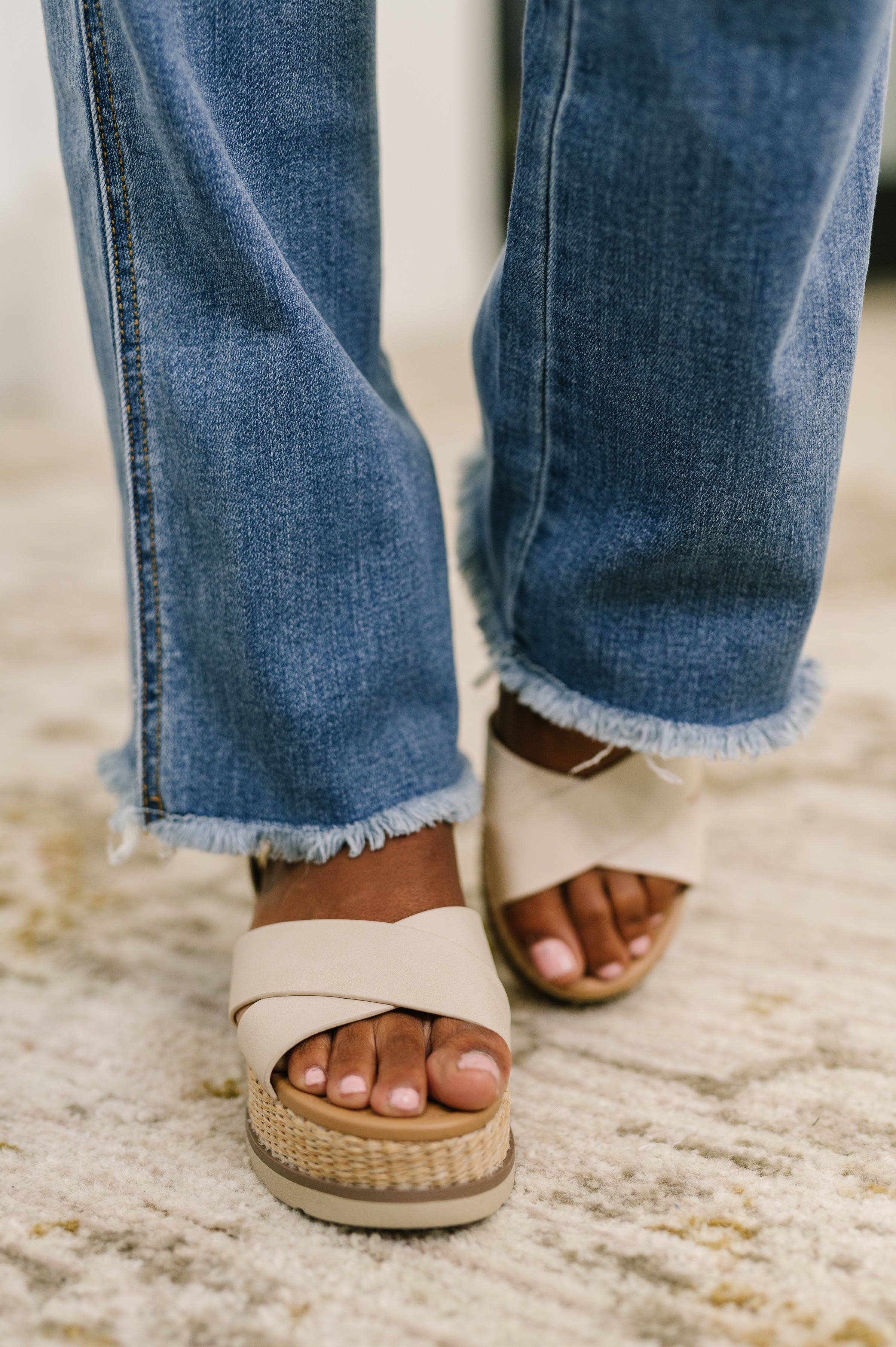 Close-up of blue high-rise bootcut jeans with raw frayed hems paired with beige crisscross platform sandals on a rug