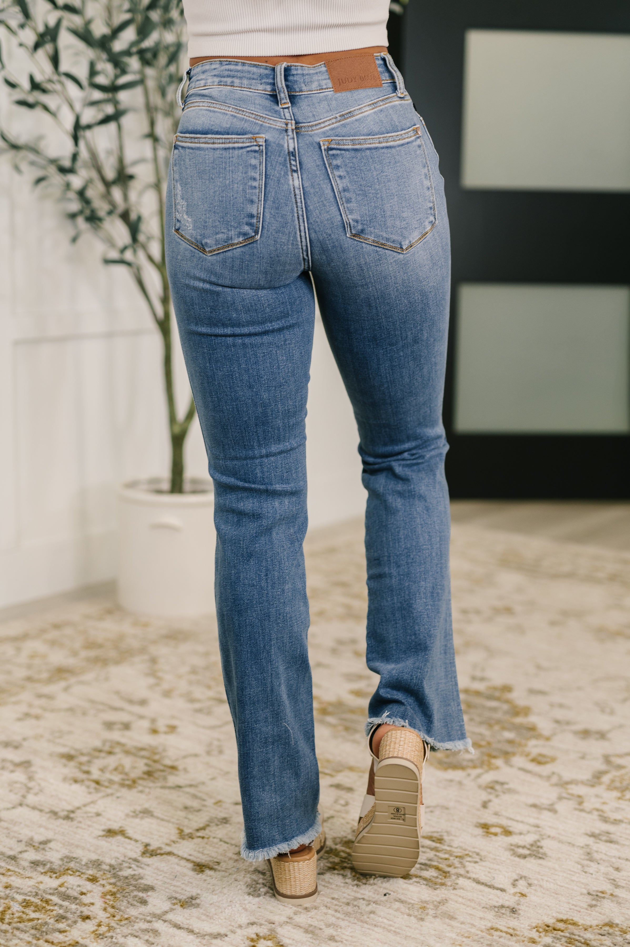 Back view of light blue high-rise jeans with subtle distressing and frayed bootcut hems worn with beige wedge sandals