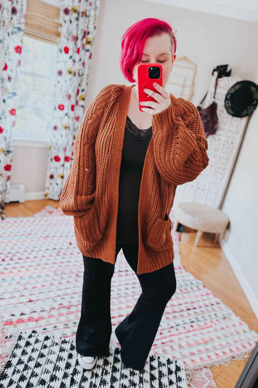 Woman with bright pink hair wearing a chestnut oversized knit cardigan with pockets over a black lace top and black pants taking a mirror selfie