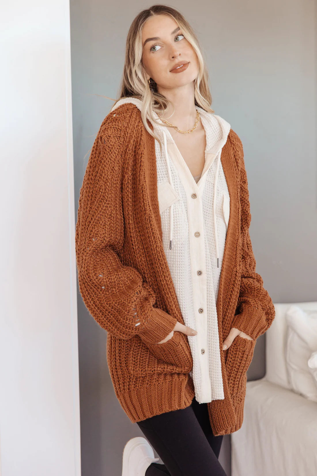 Woman wearing a chestnut knit cardigan with balloon sleeves and pockets over a cream waffle-knit hoodie indoors