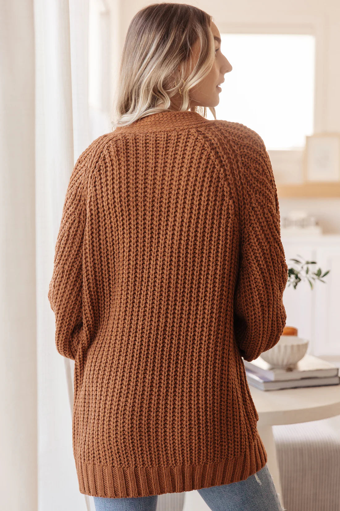 Back view of a woman wearing a chestnut chunky knit cardigan with a textured pattern and ribbed hem indoors