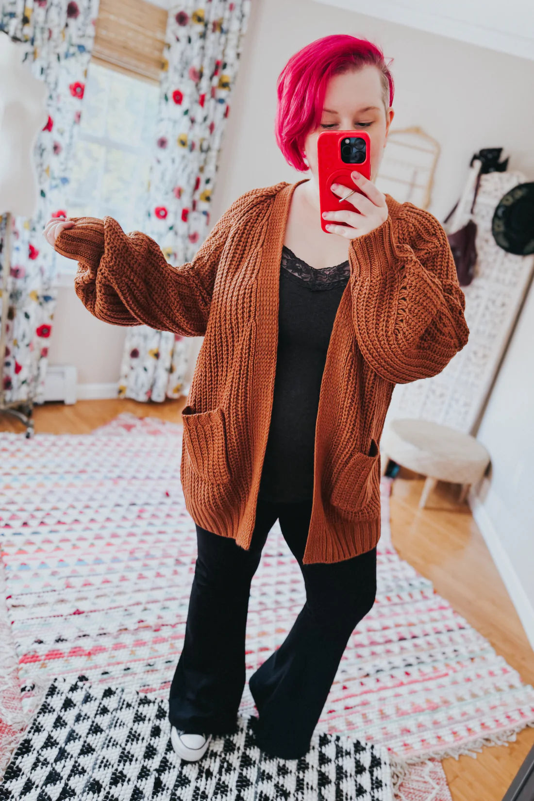 Woman wearing a chestnut chunky knit cardigan with large pockets over a black lace-trimmed top indoors taking a mirror selfie