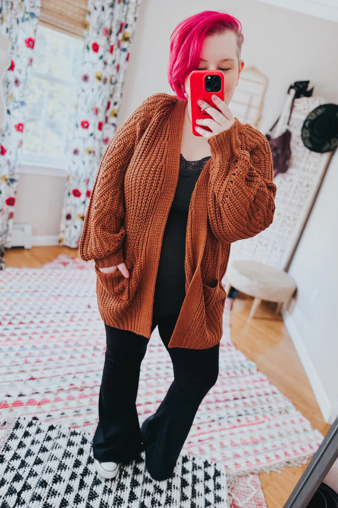 Woman with bright pink hair wearing a chestnut oversized knit cardigan with pockets over black lace top and pants indoors