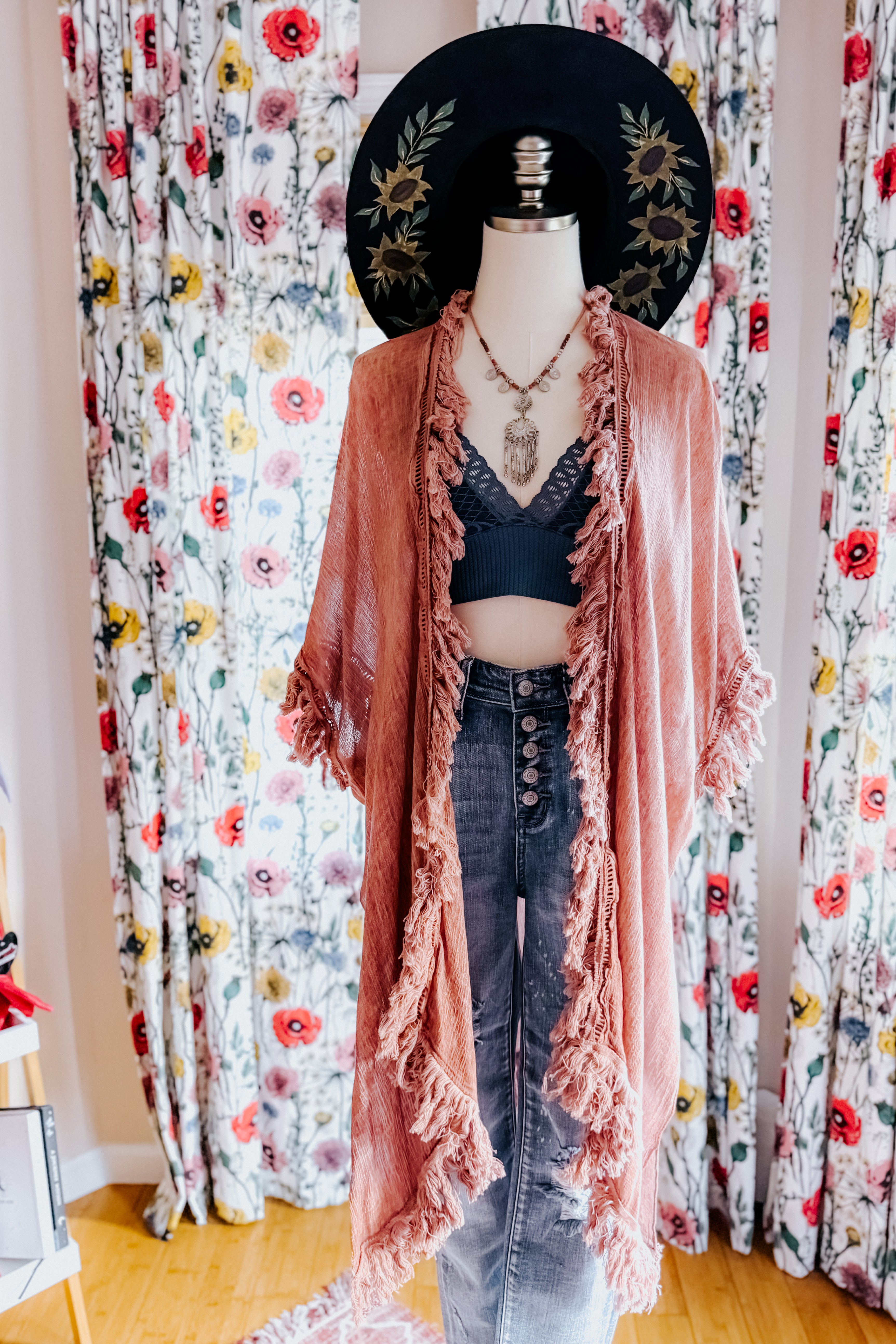 Mannequin styled with sunbleached rose fringe duster, black lace bralette, high-waisted jeans, layered necklace, and wide-brim hat indoors