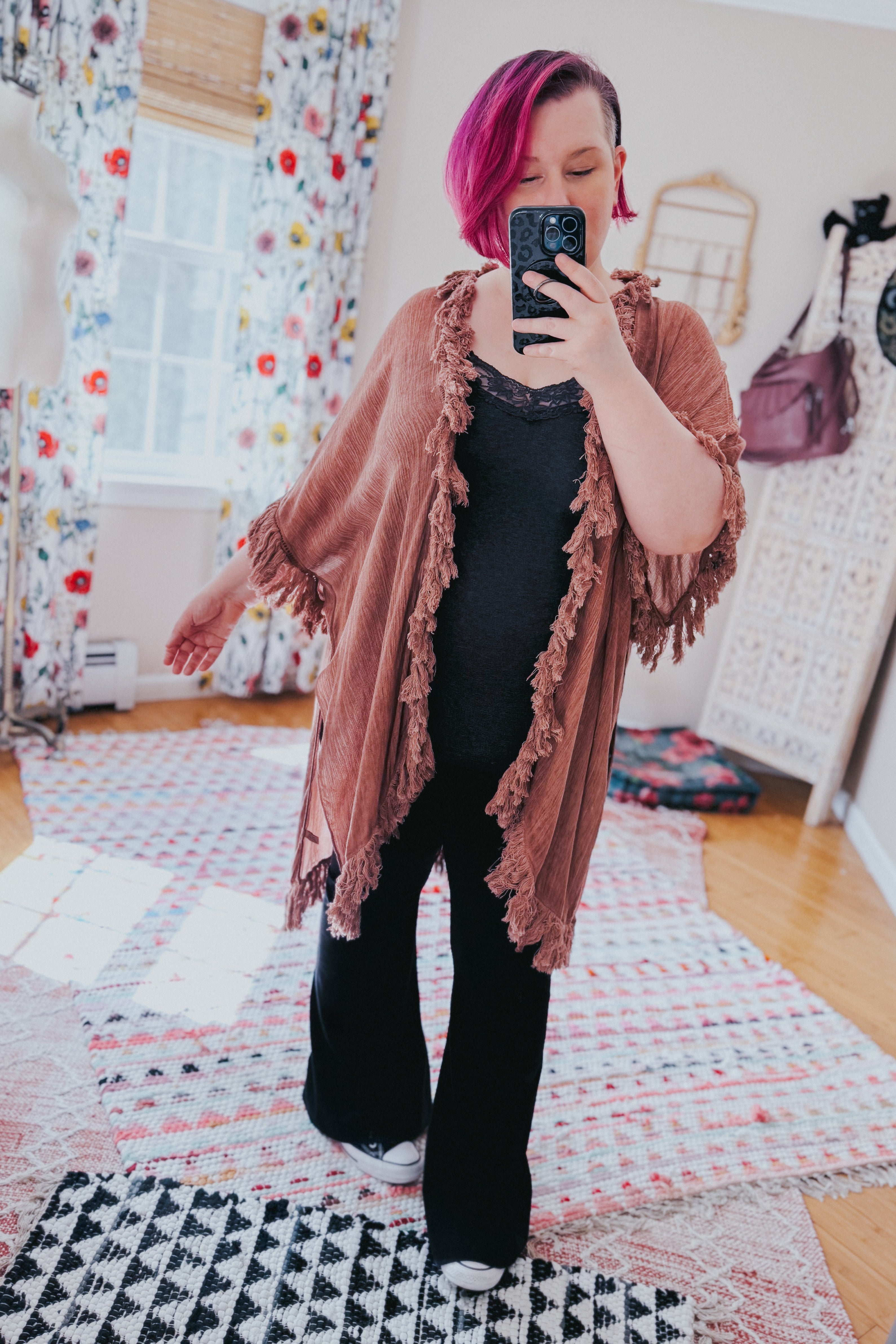 Woman with magenta hair wearing a rose-colored fringed duster over black outfit in a bright boho-style room