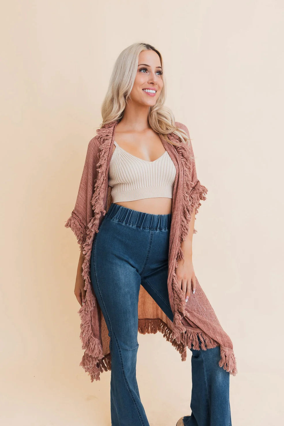 Blonde woman wearing a rose-colored fringe duster over a cream crop top and high-waisted blue jeans smiling