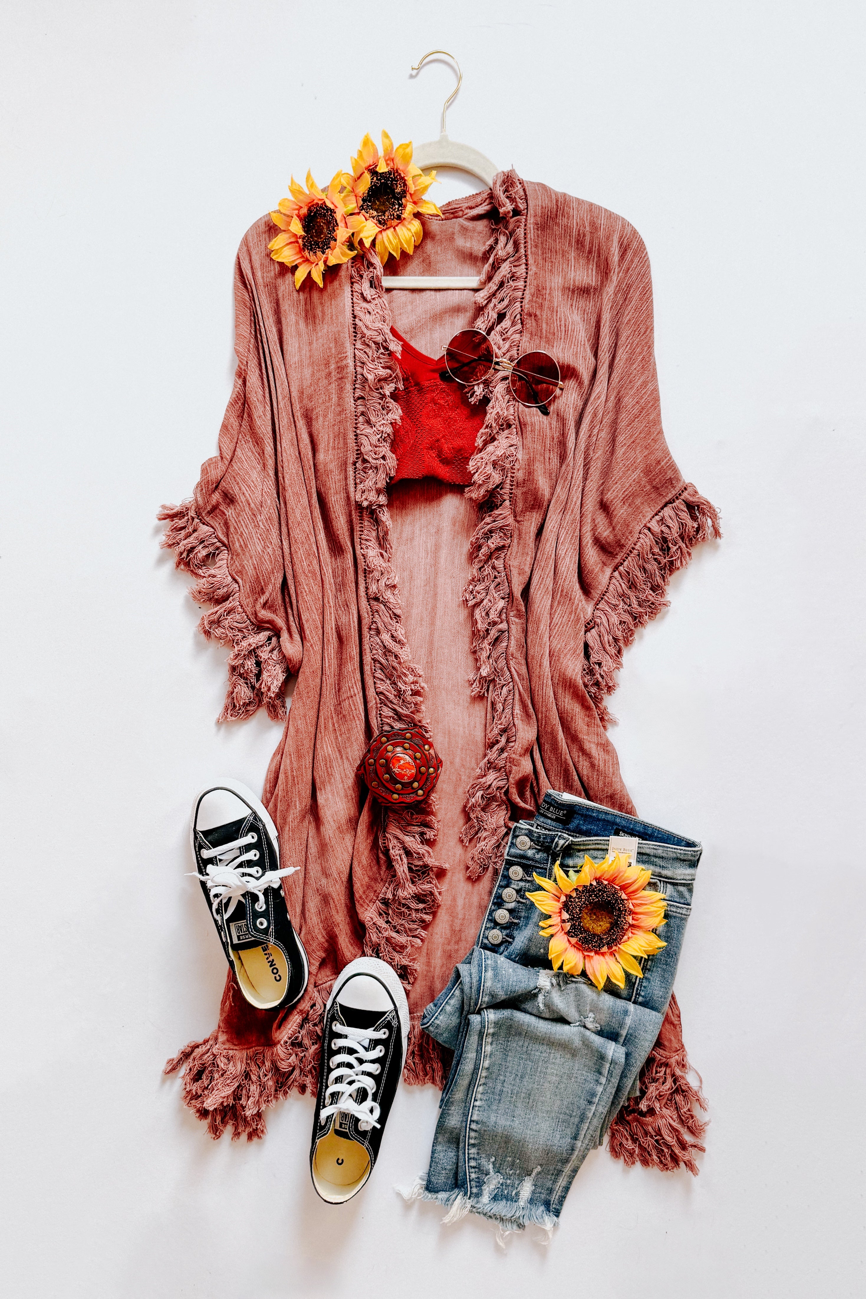 Rose-colored fringed duster layered over a red top, paired with distressed jeans, black sneakers, and sunflower accents