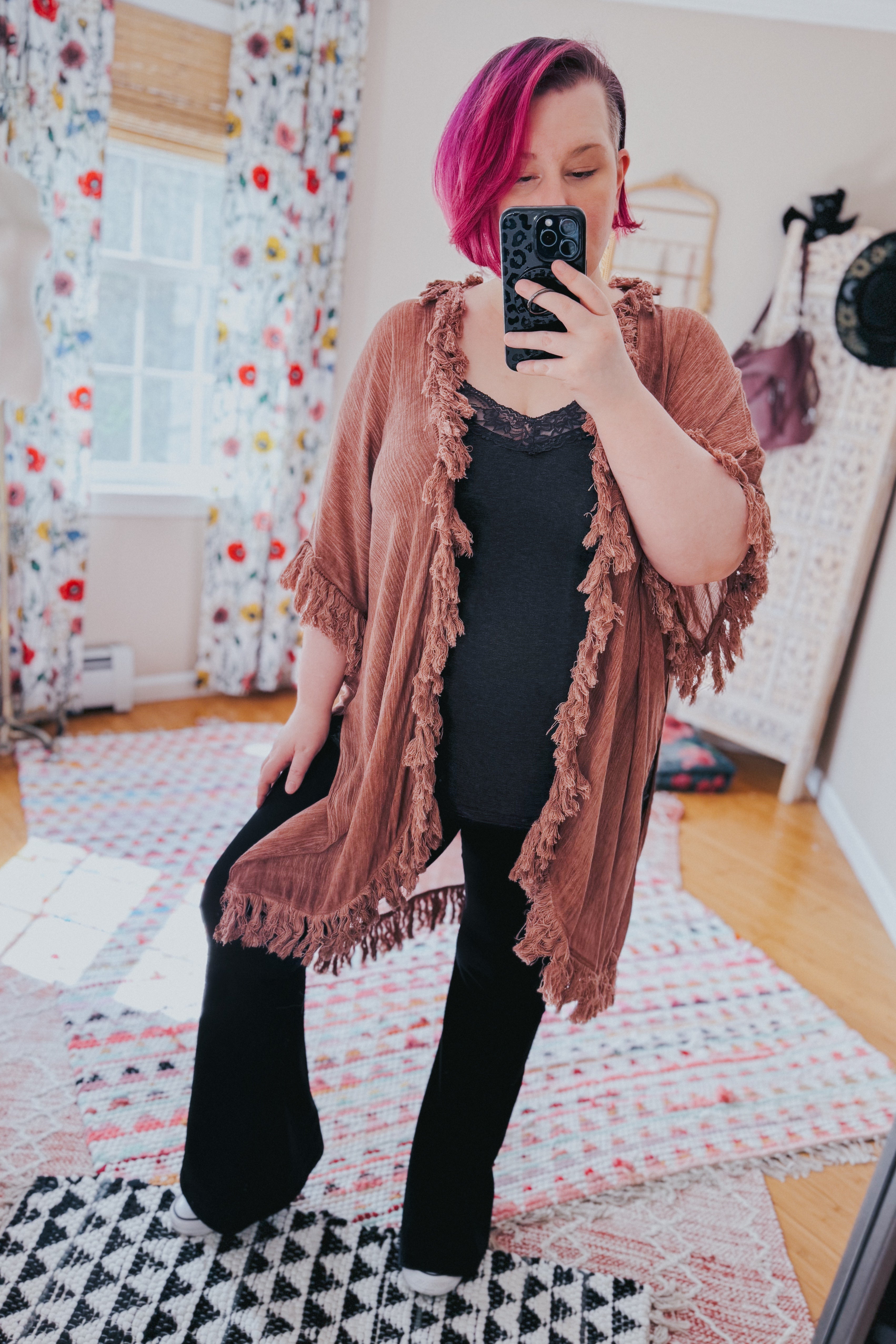 Woman wearing a rose-colored fringed duster layered over black top and pants, taking a mirror selfie in a boho room