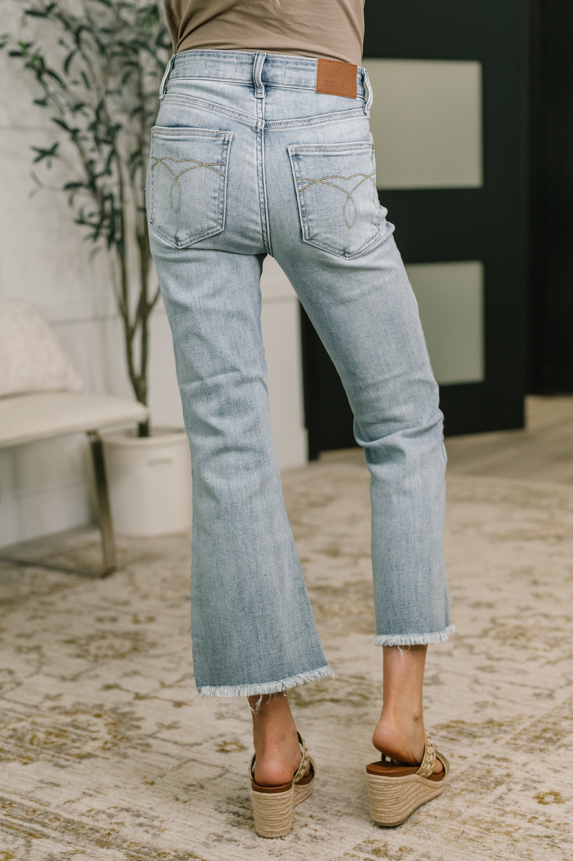 Rear view of light blue mid rise flare jeans with frayed hems worn with tan wedge sandals indoors