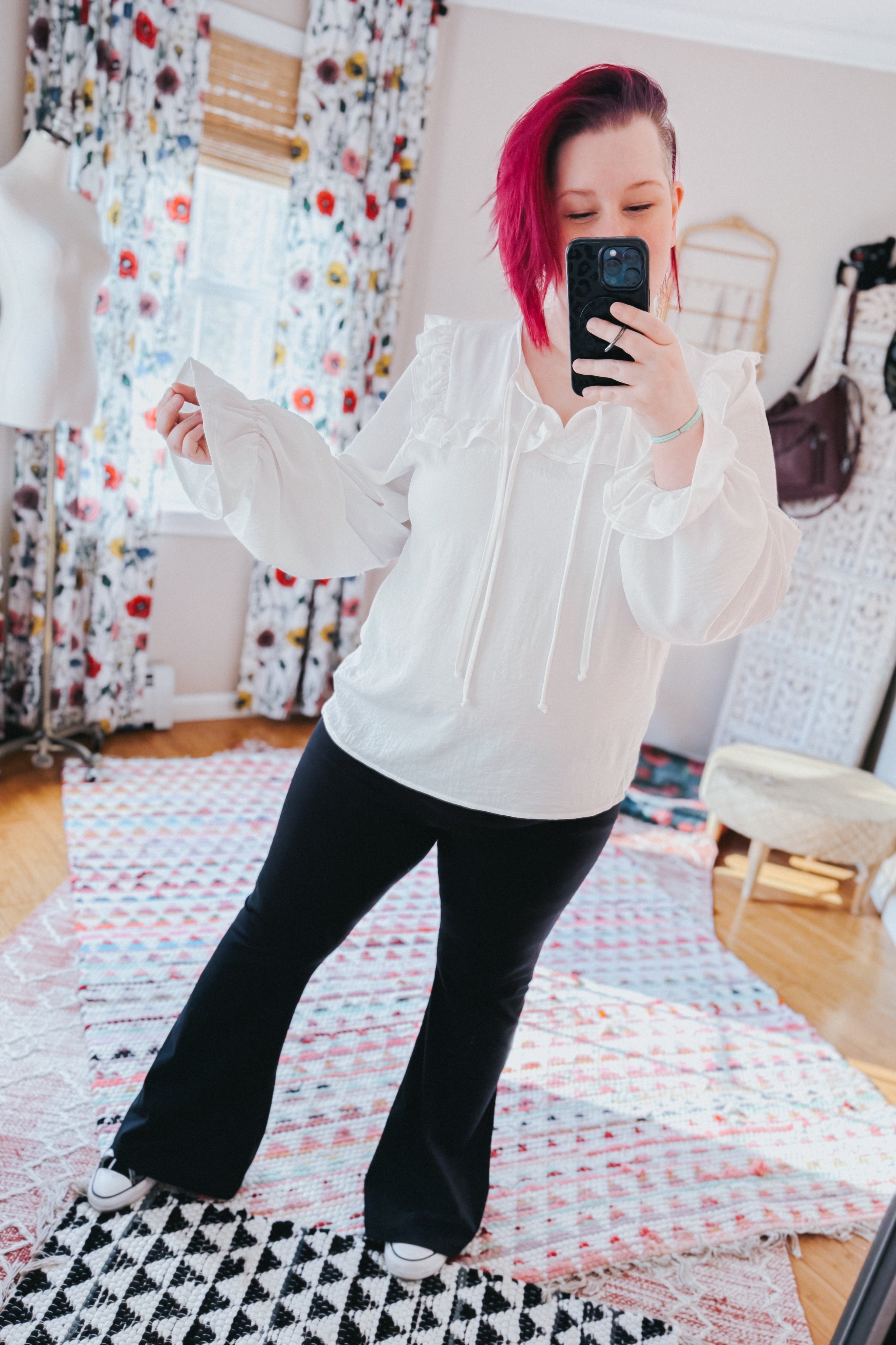 Woman taking a mirror selfie wearing a white blouse with voluminous bubble sleeves and ruffle details, paired with black flared pants indoors.