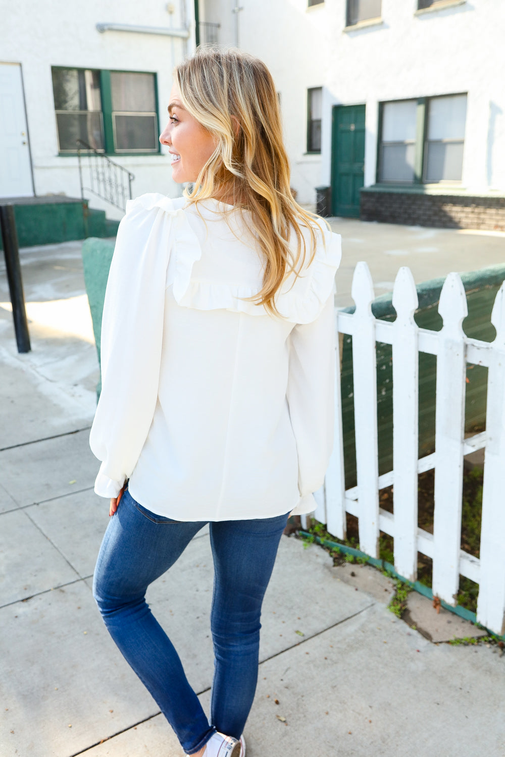 Woman with blonde hair wearing a white blouse featuring bubble sleeves and ruffle details on the back, paired with blue jeans, standing outdoors near a white picket fence.