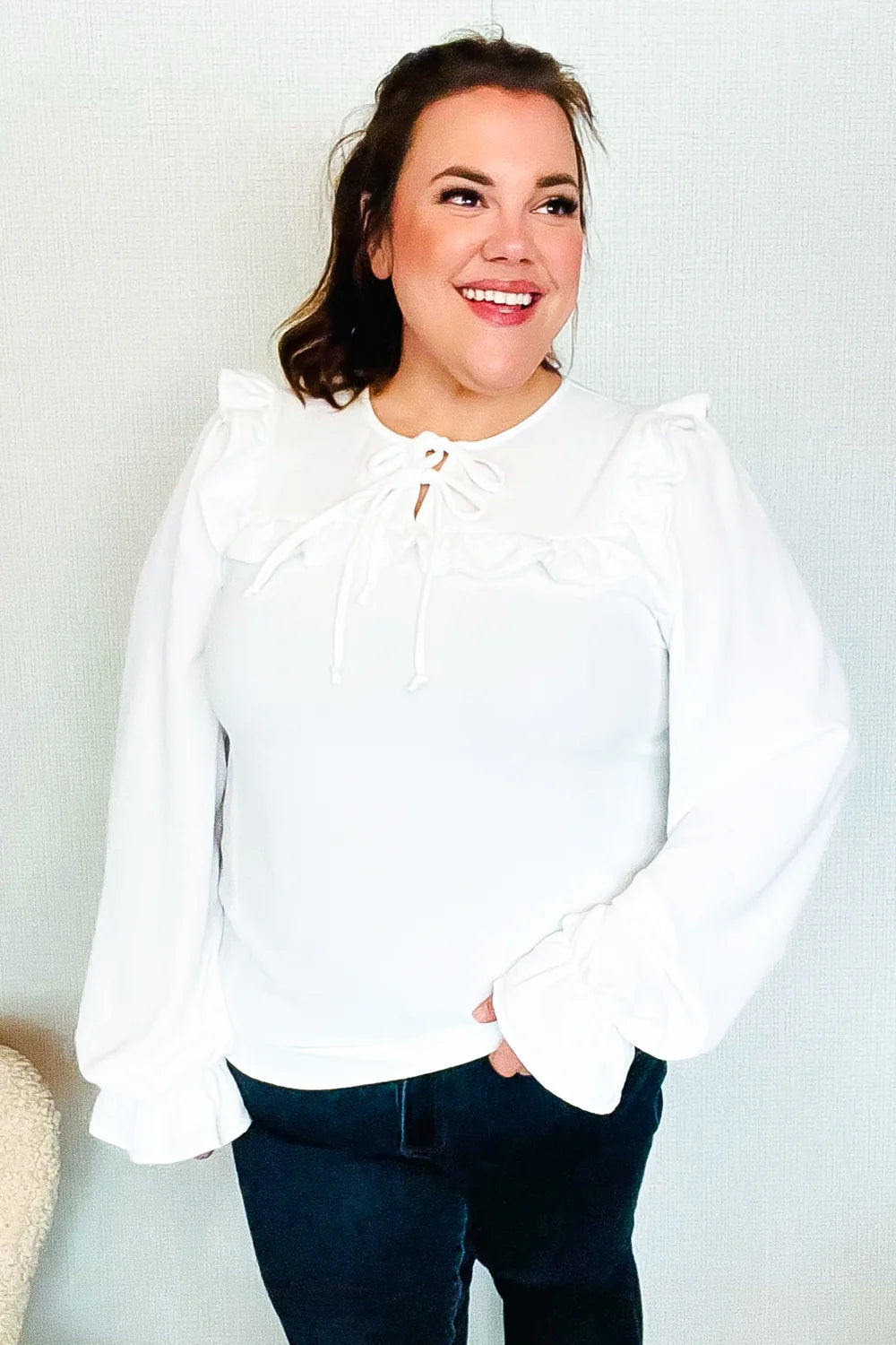 Smiling woman wearing a white blouse with puffed sleeves, ruffle trim along the chest, and a tie detail at the neckline, paired with dark jeans.