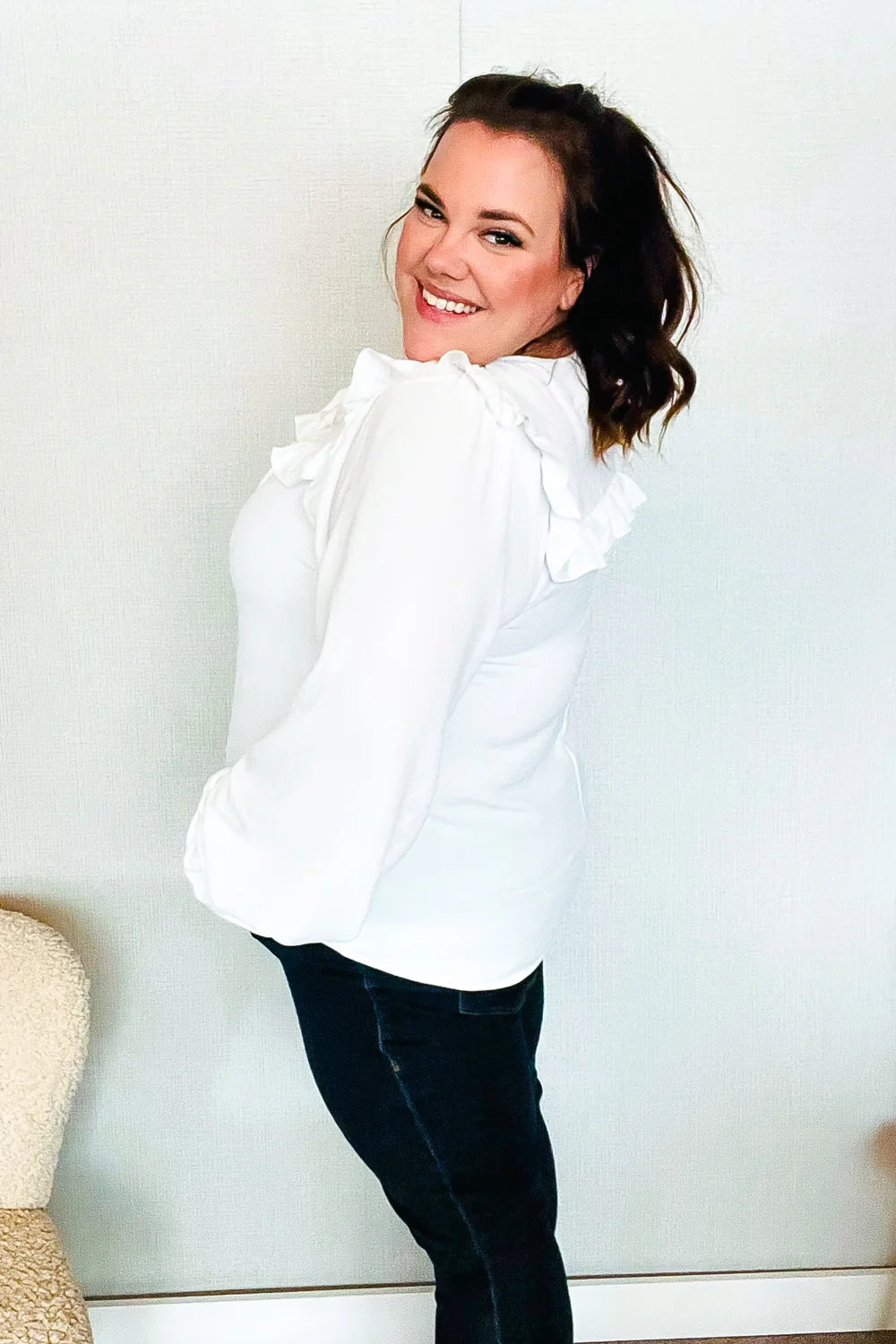 Smiling woman wearing a white blouse with ruffled shoulder details and voluminous sleeves, paired with dark fitted pants indoors.