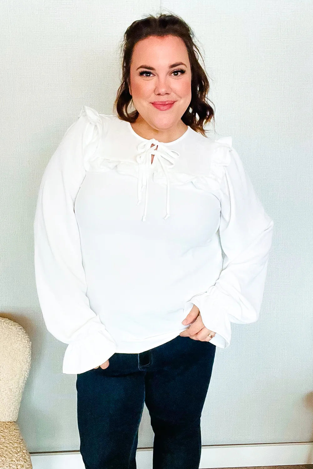 Smiling woman wearing a white blouse with delicate ruffle accents and a tie detail at the neckline, paired with dark jeans indoors.