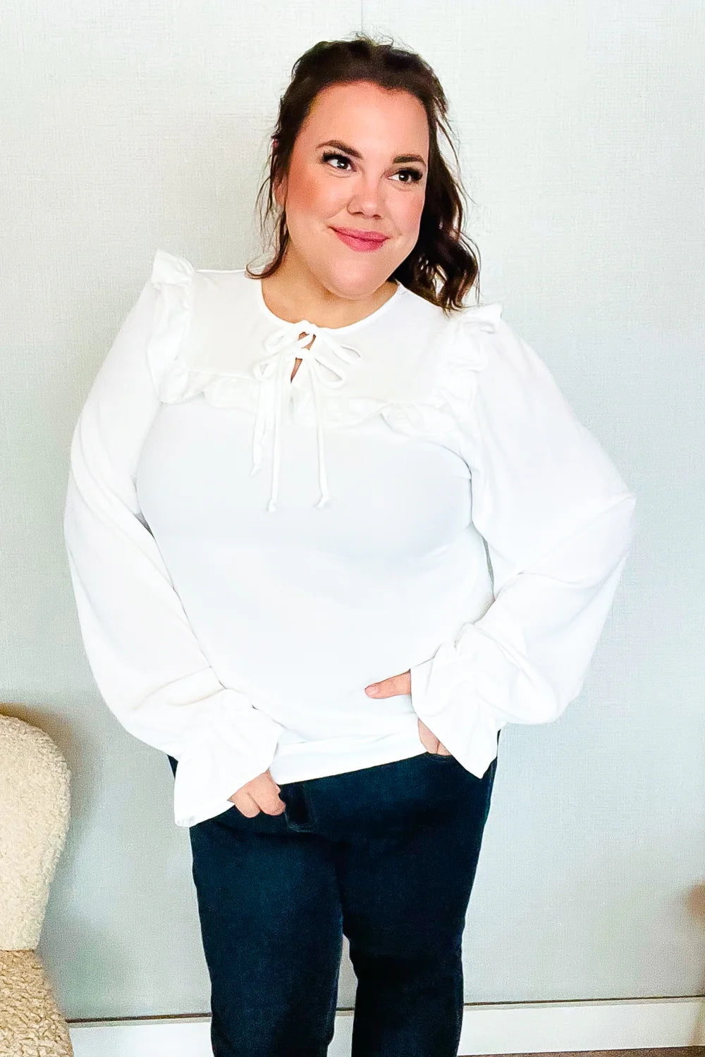 Woman wearing a white blouse with bubble sleeves, ruffle detailing on the shoulders, and a front tie neckline, paired with dark pants indoors.