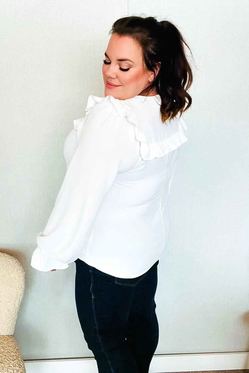 Woman wearing a white blouse with puffed sleeves and a ruffled yoke detail, paired with dark pants, standing indoors facing sideways.