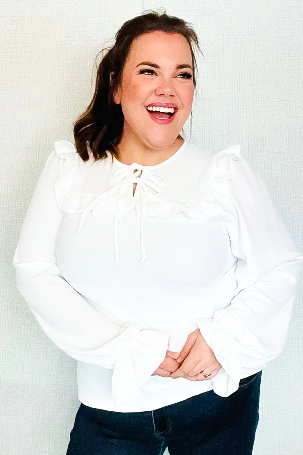 Woman wearing a white blouse with bubble sleeves, ruffle trim at the chest, and tie detail at the neckline, smiling indoors.