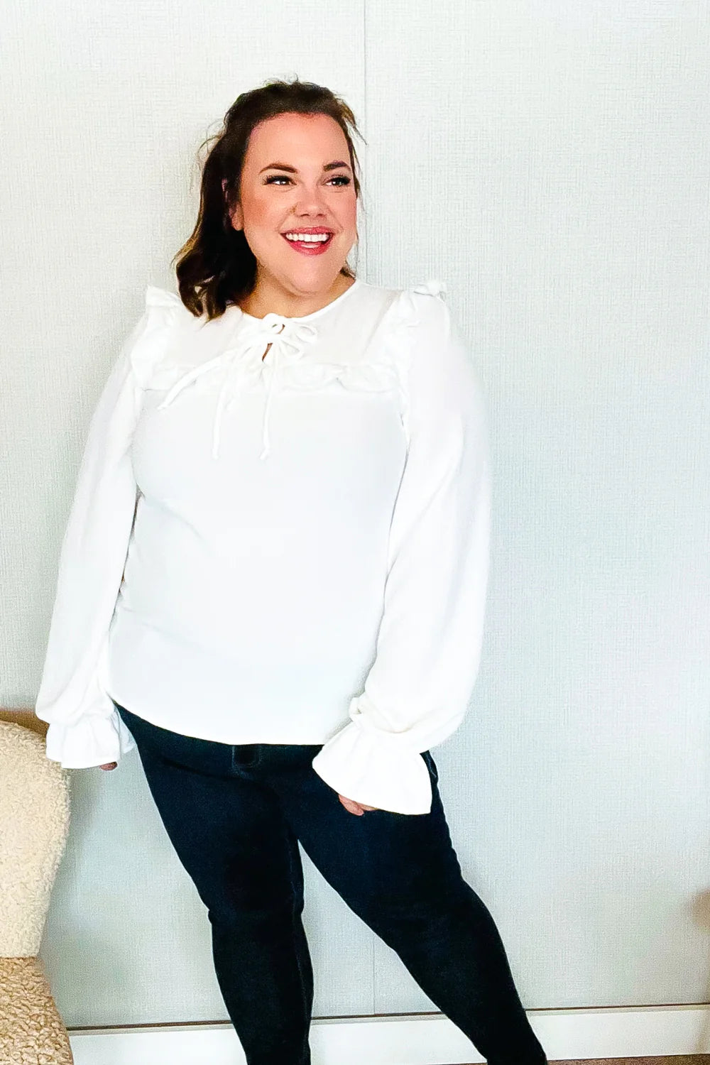 Smiling woman wearing a white blouse with long puffed sleeves, ruffle details near the neckline, and a tie closure, paired with dark pants indoors.
