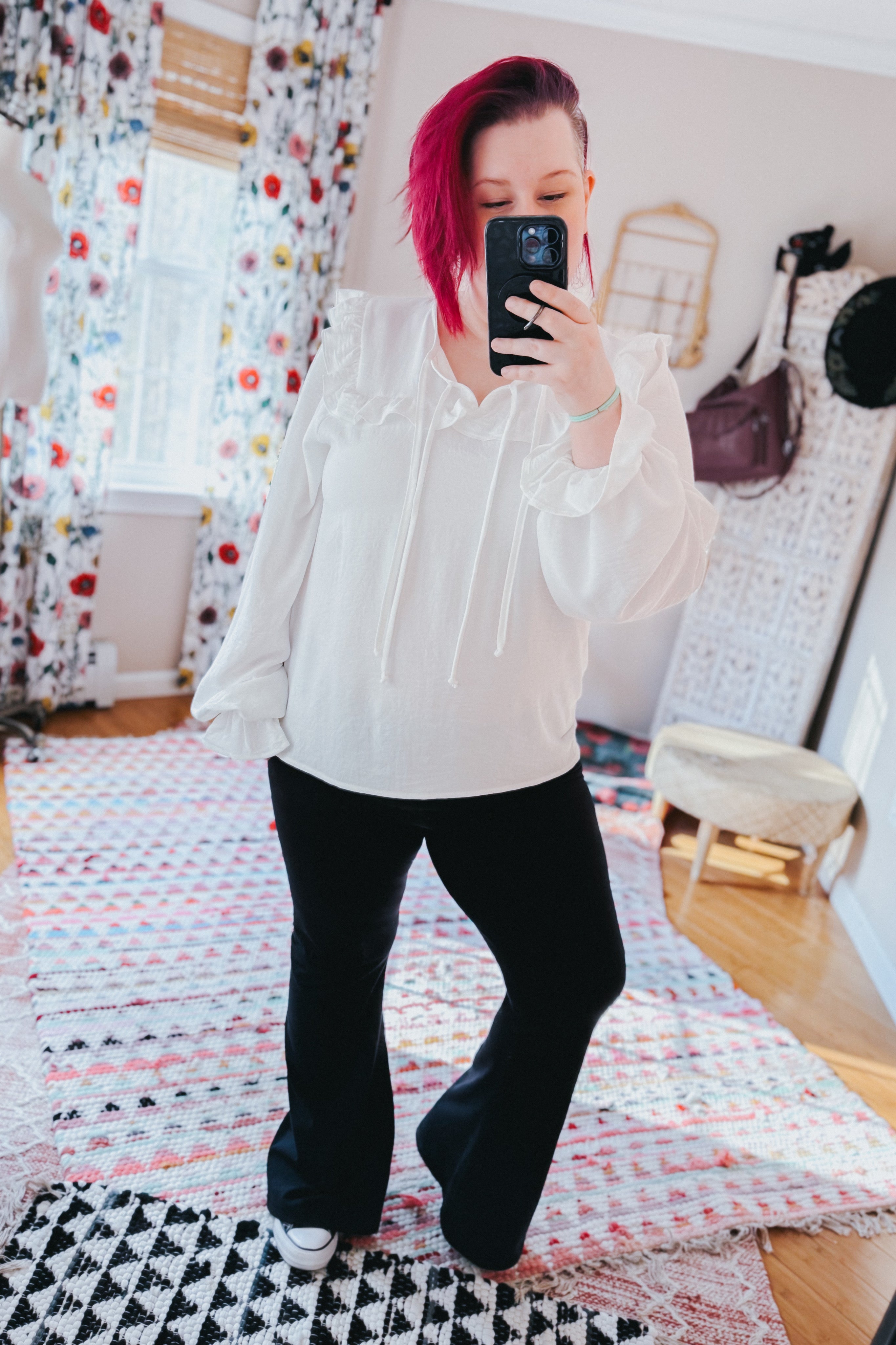 Woman taking a mirror selfie wearing a white blouse with ruffled neckline and billowy sleeves, paired with black flared pants indoors.