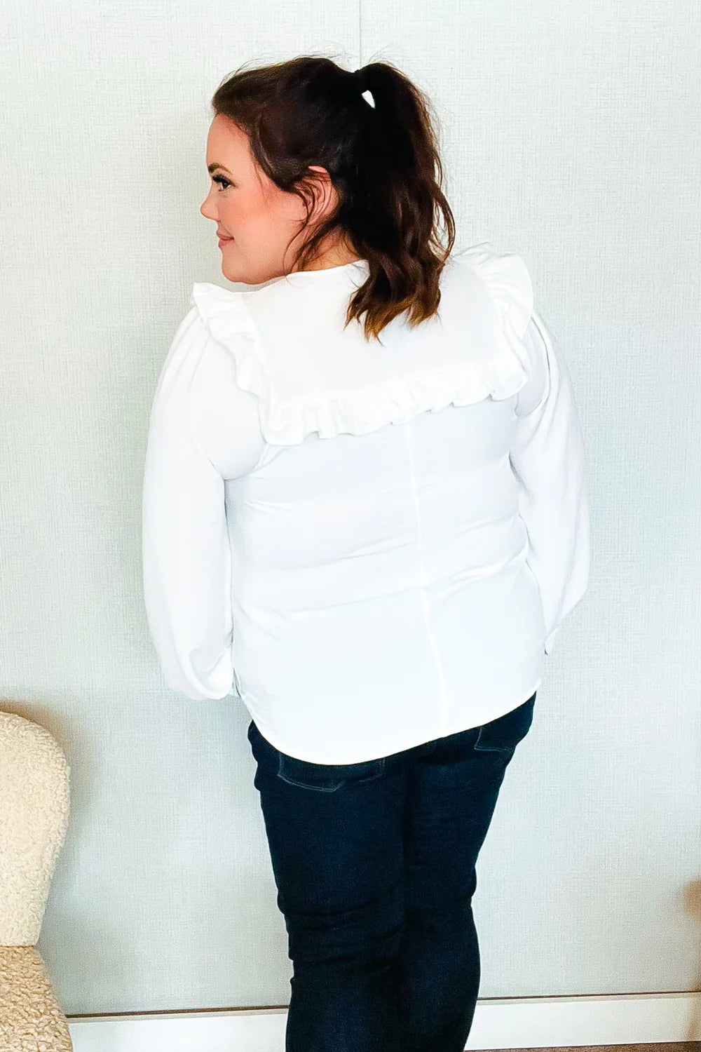 Woman wearing a white blouse with bubble sleeves and a wide ruffled yoke across the back, paired with dark fitted jeans, standing indoors.