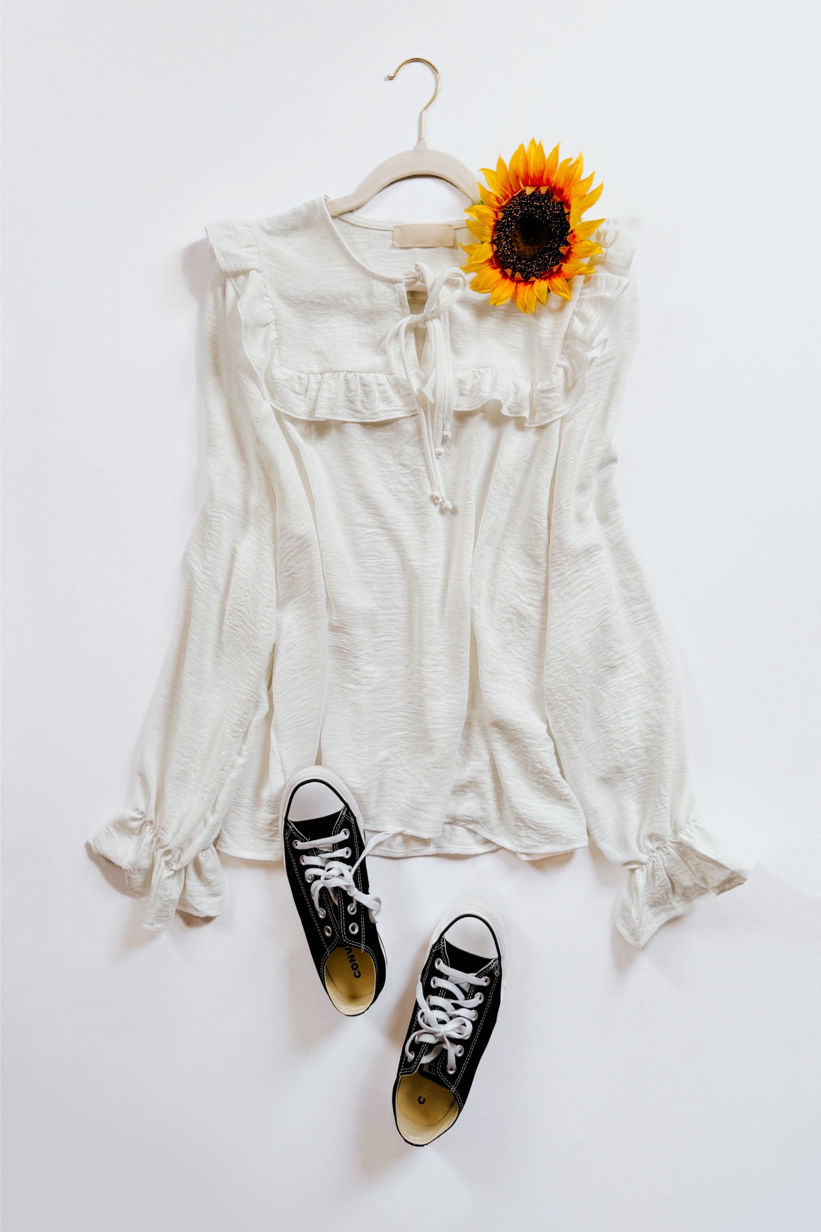 White blouse with bubble sleeves, ruffled yoke, and tie neckline displayed on a hanger with a sunflower and black canvas sneakers below.