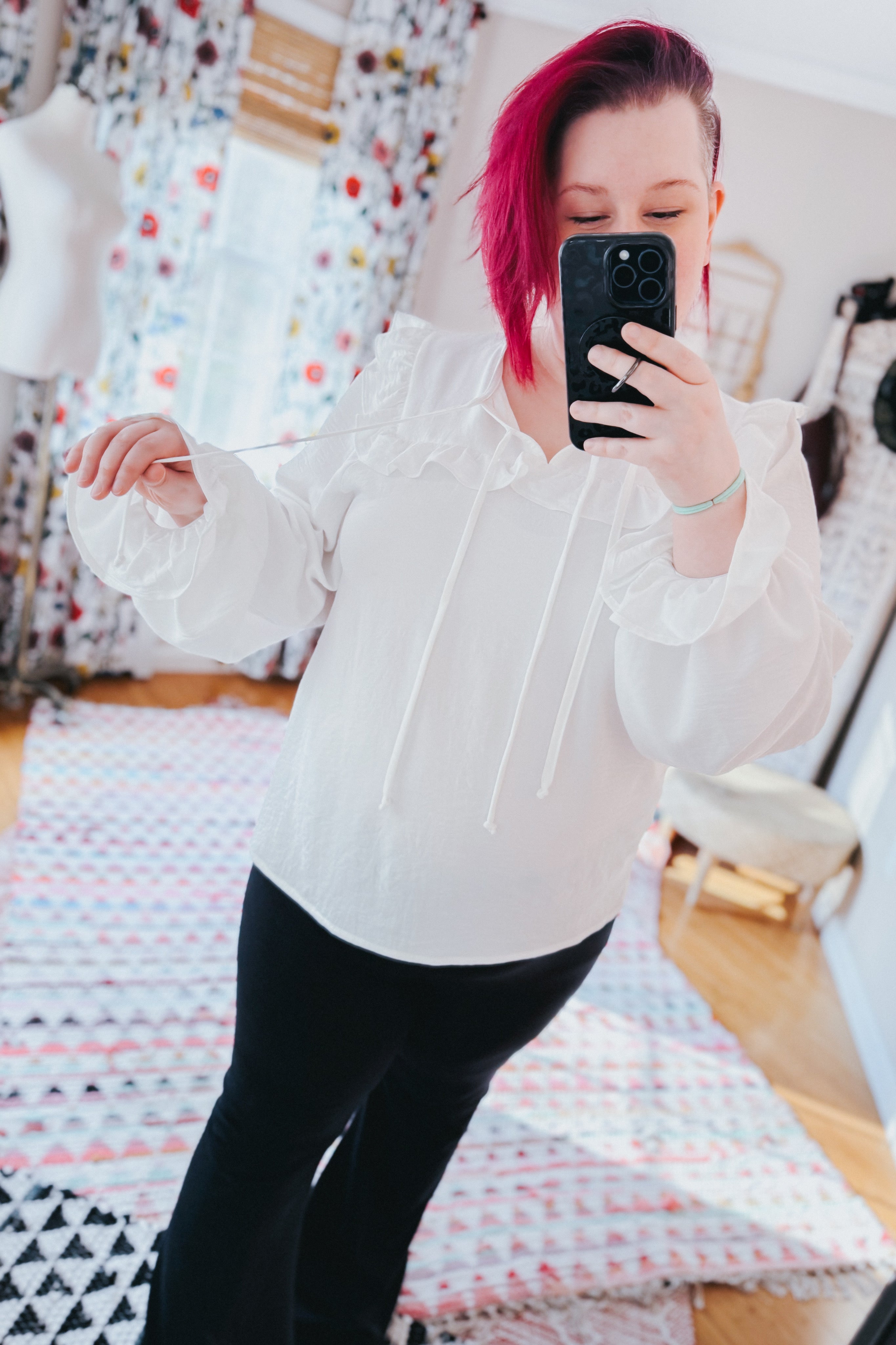Woman taking a mirror selfie wearing a white blouse with long ruffled sleeves and a tie neckline, paired with black pants indoors.