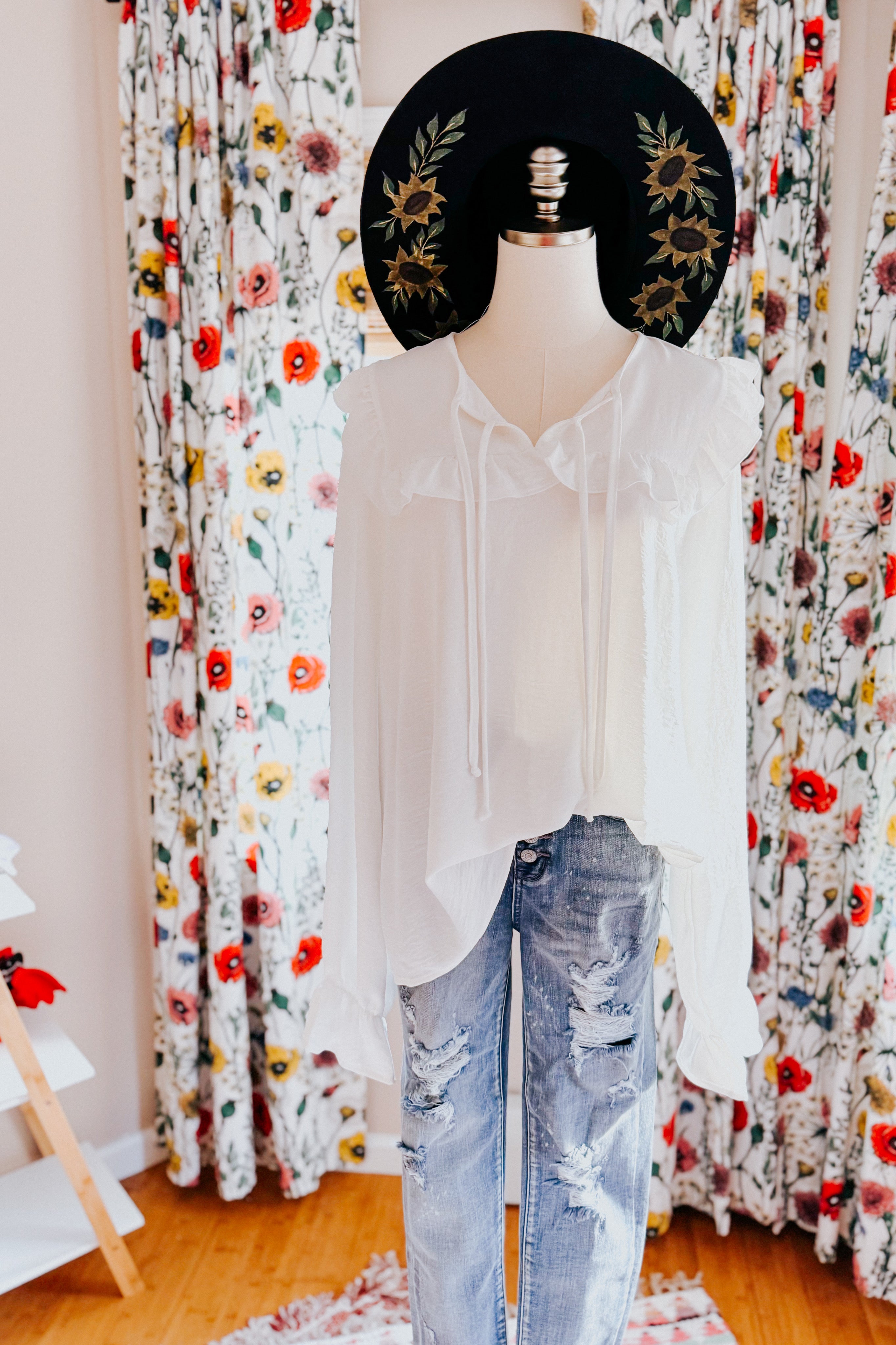 White bubble sleeve blouse with ruffled collar and tie detail styled on a mannequin with distressed blue jeans and a black wide-brim hat decorated with sunflowers.