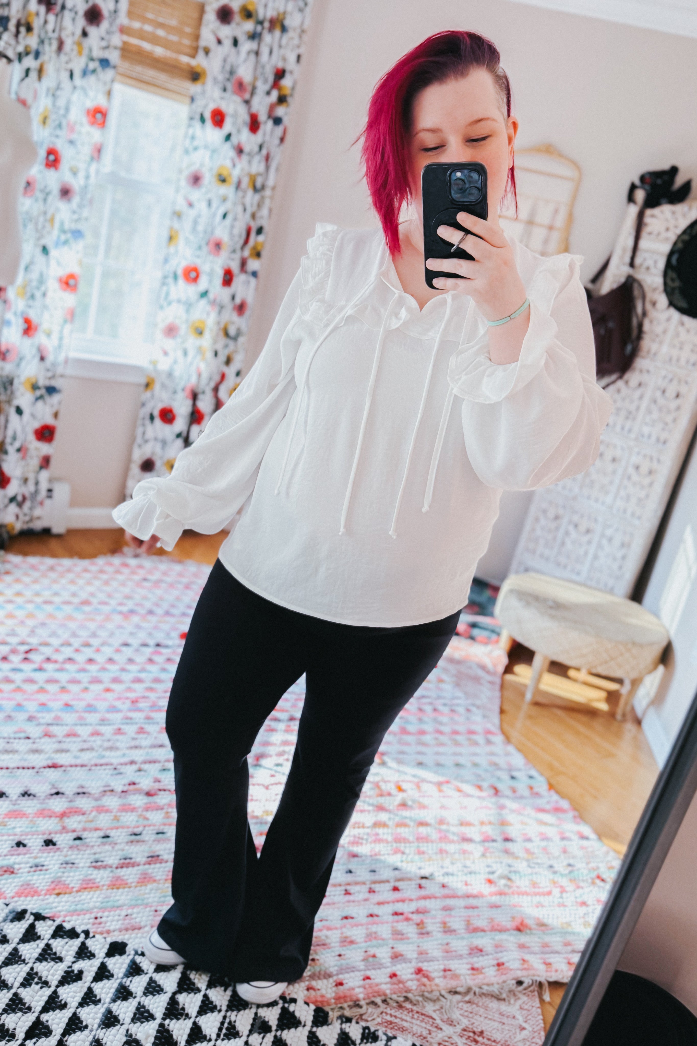 Woman taking a mirror selfie wearing a white blouse with ruffled shoulders and long bubble sleeves, paired with black pants indoors.