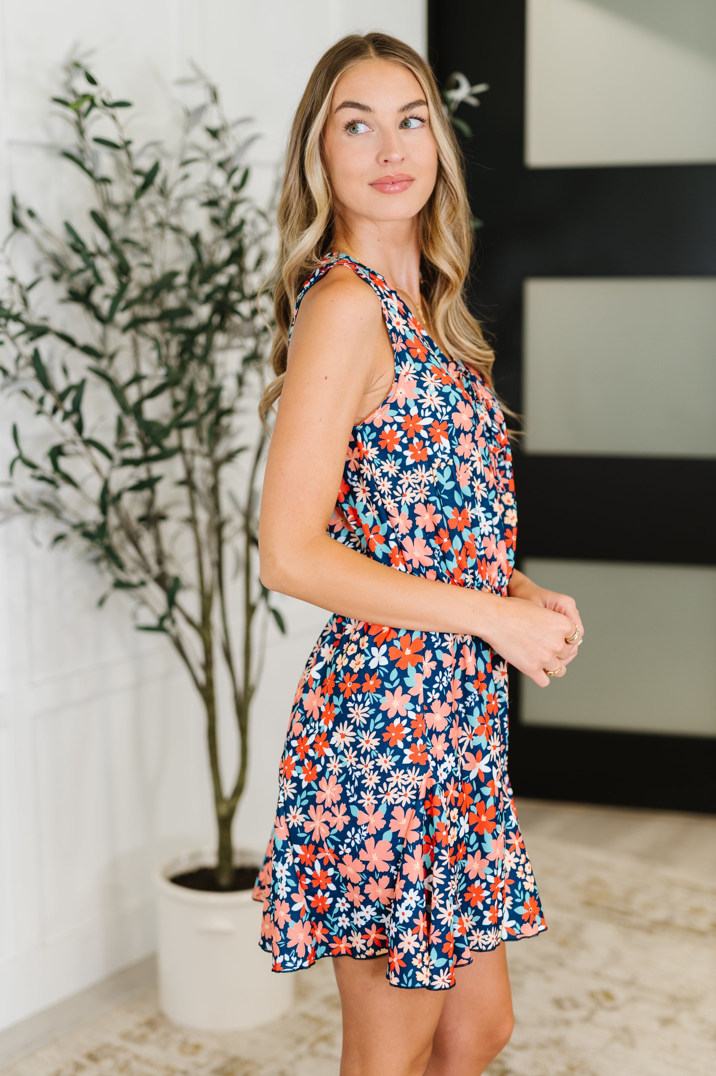 Woman wearing sleeveless navy romper with orange and teal floral print and neck tie detail in indoor setting