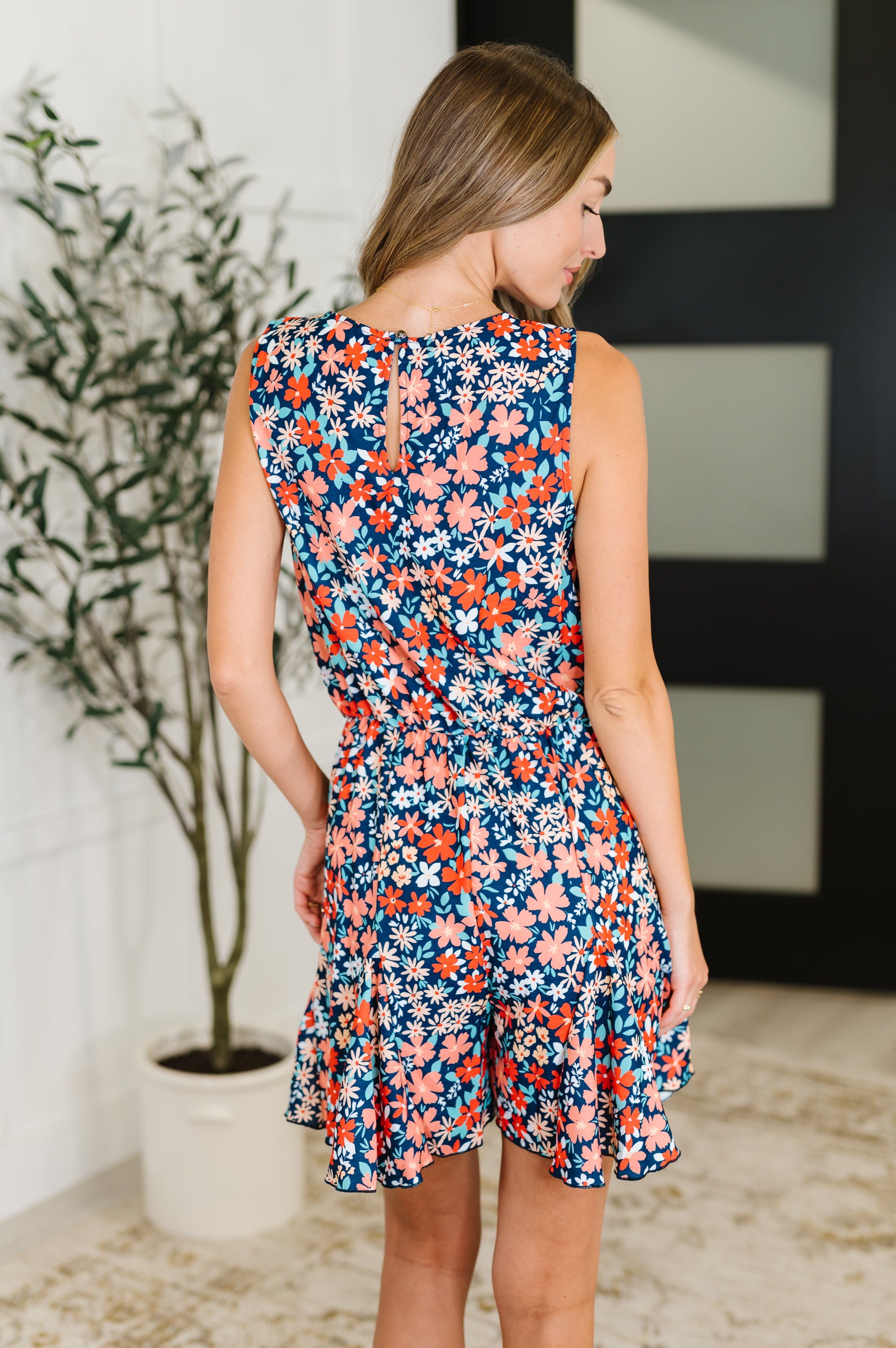 Back view of woman wearing sleeveless navy romper with colorful floral print and ruffle hem in bright indoor space
