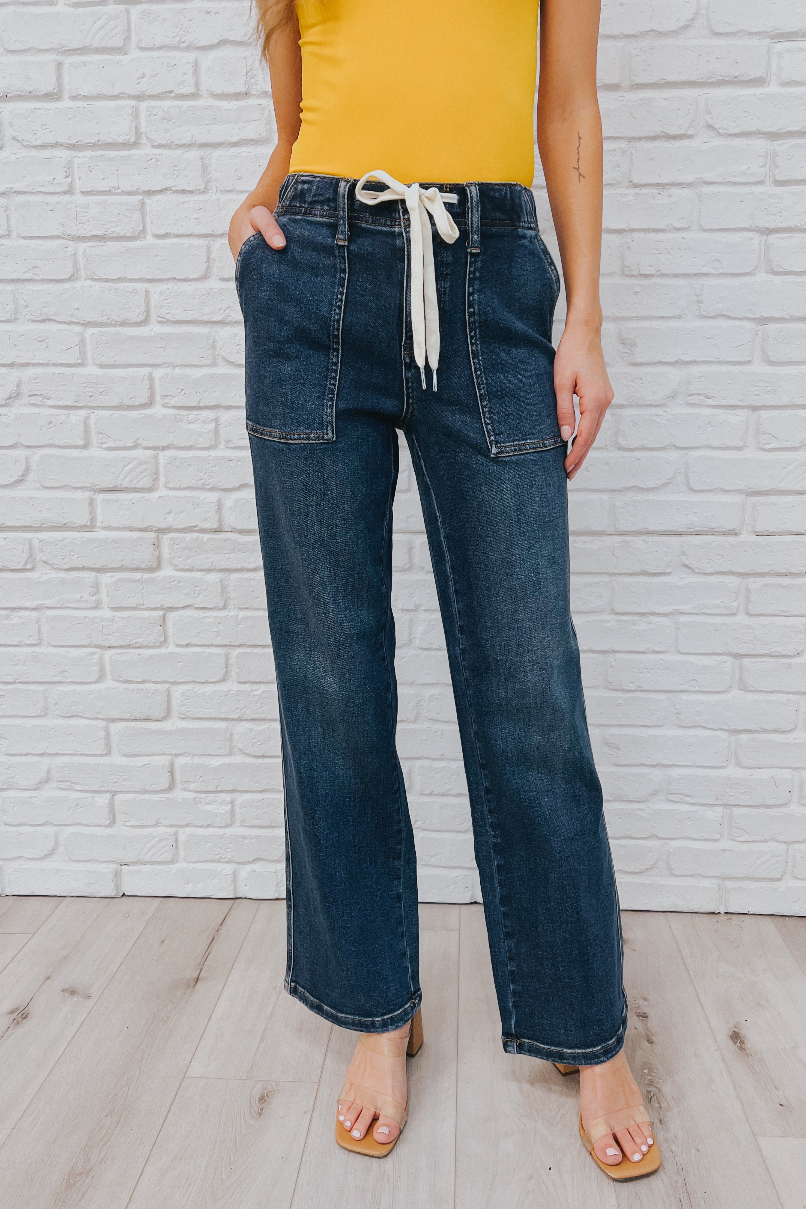 Woman wearing high-rise wide leg blue jeans with large front pockets and a white drawstring belt, paired with a mustard yellow sleeveless top.