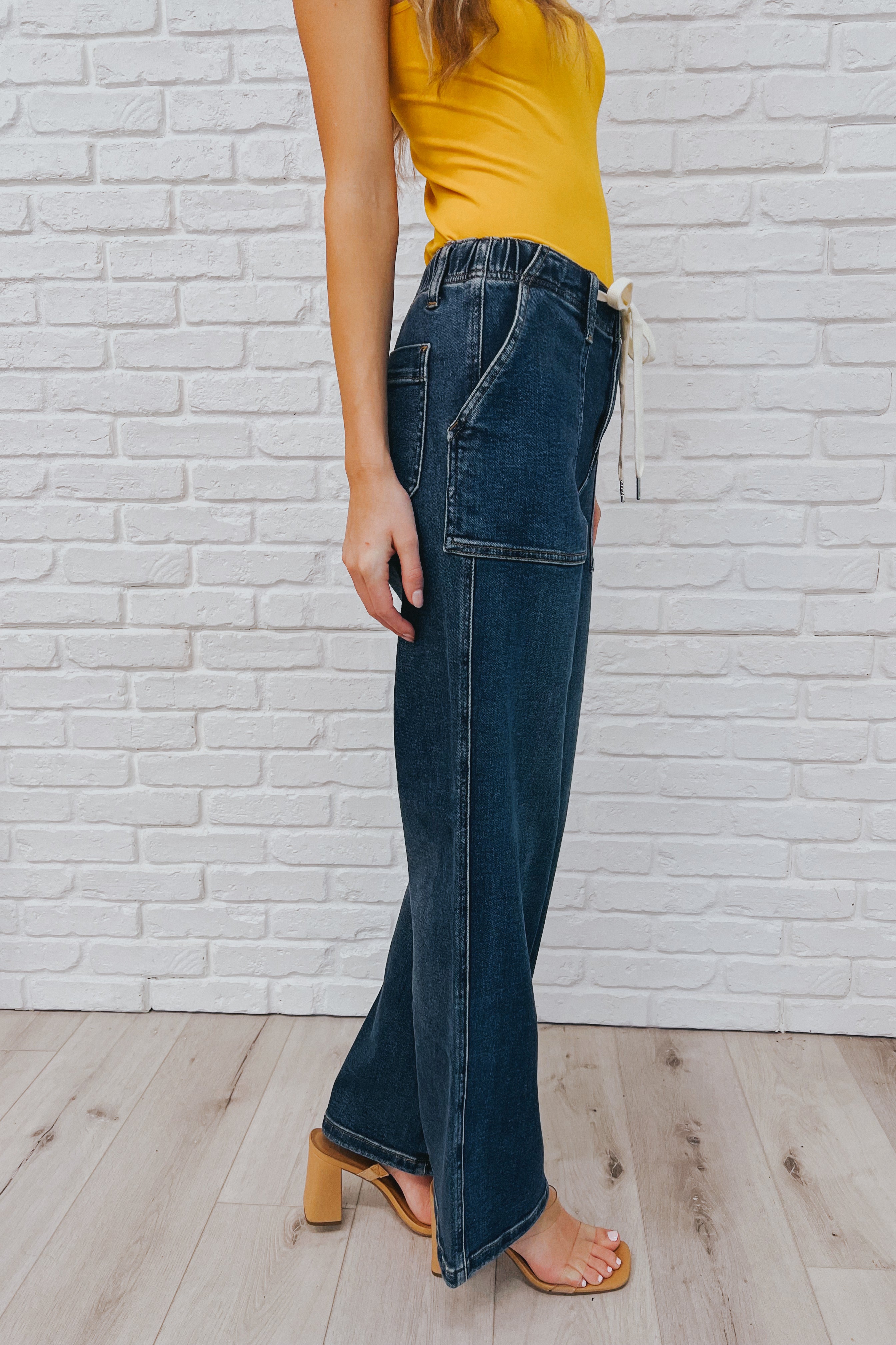 Side view of a woman wearing dark blue high-rise wide-leg jeans with large patch pockets and a white drawstring belt, paired with a mustard yellow sleeveless top and tan heeled sandals.