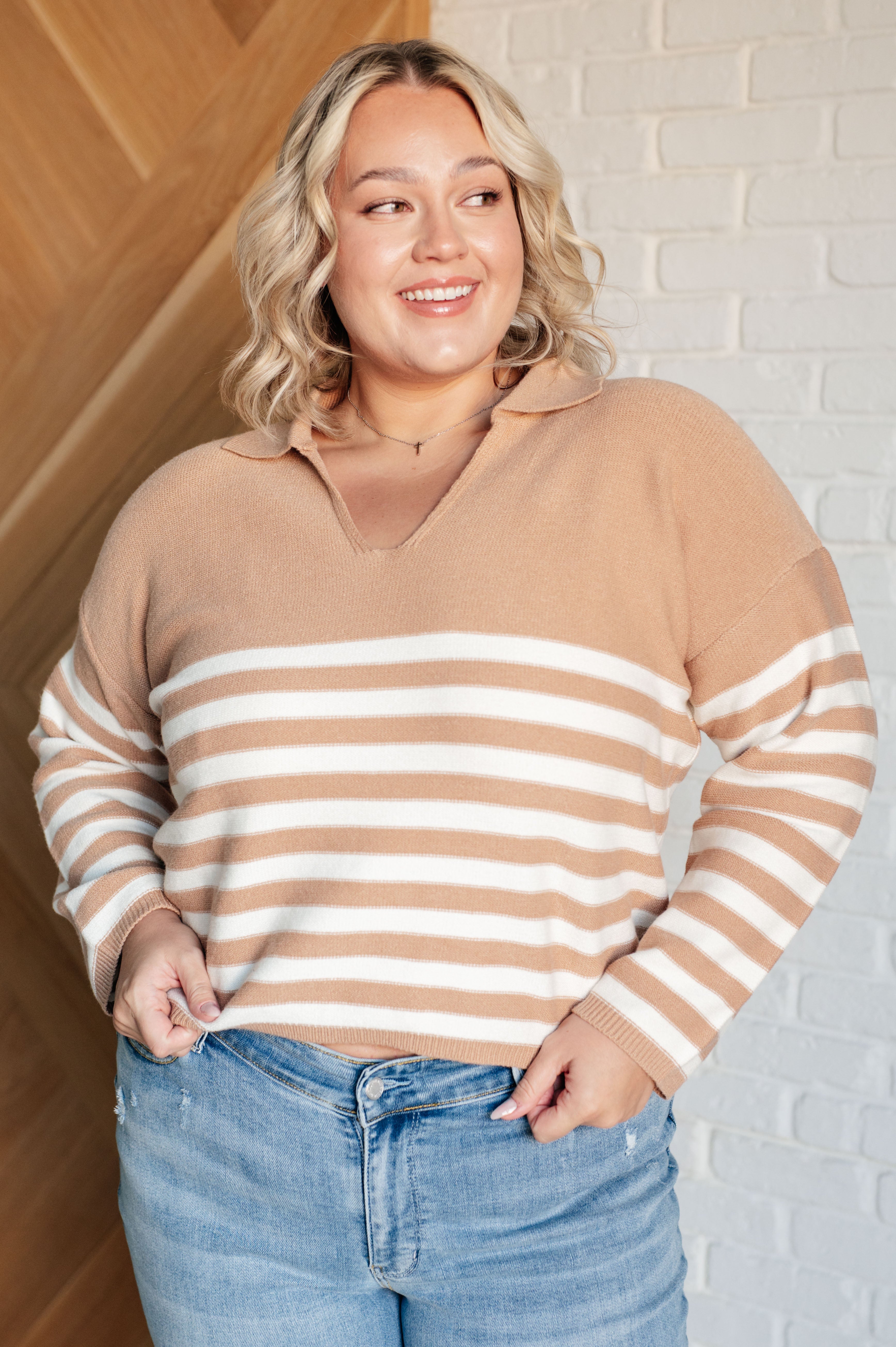 Smiling woman wearing a mocha and white striped sweater with a collar and light wash jeans, standing indoors against a white brick wall.