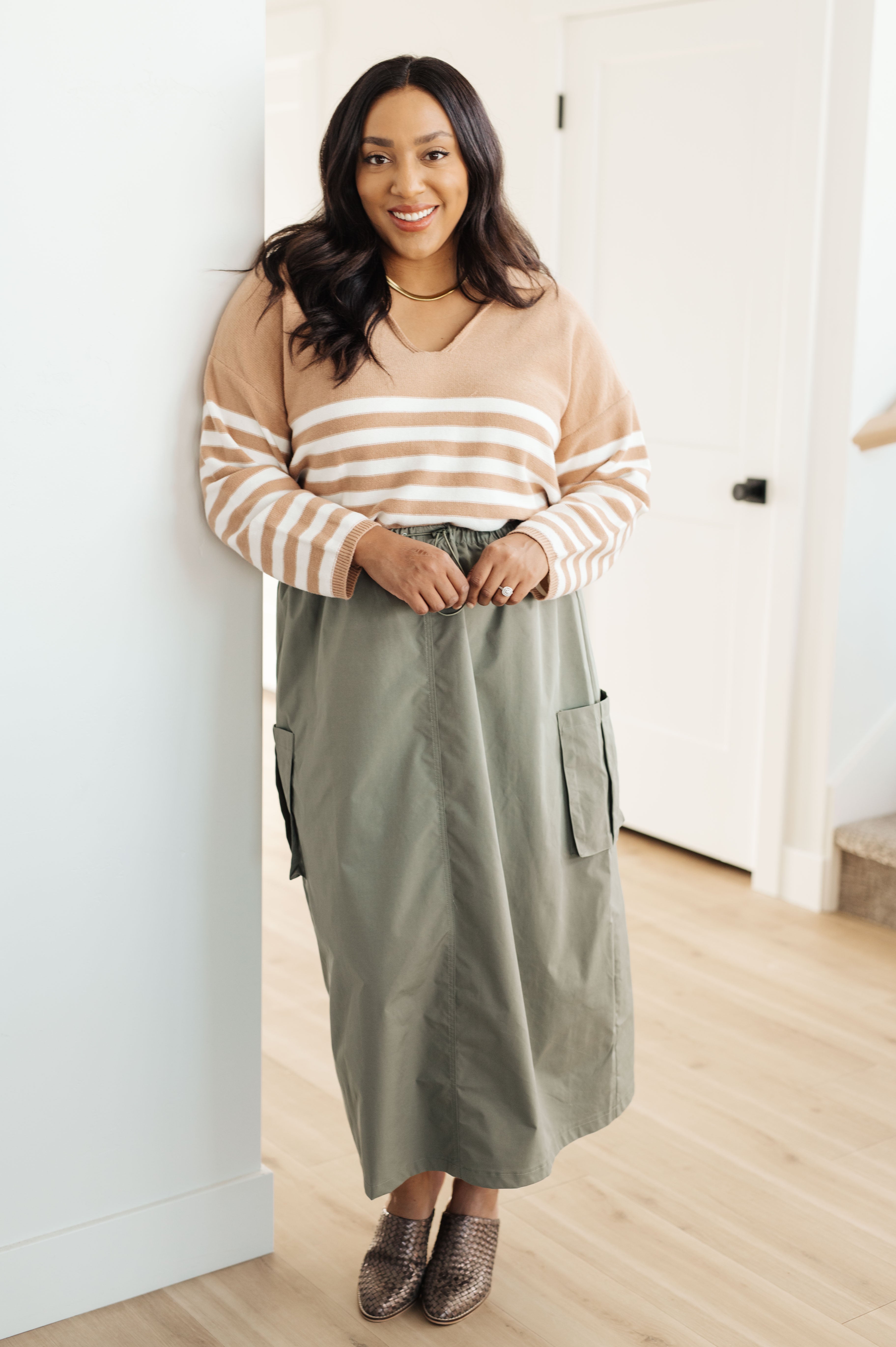 Woman wearing a mocha striped sweater with a relaxed fit and a v-neck, paired with an olive green midi skirt with pockets and patterned ankle boots, standing indoors.