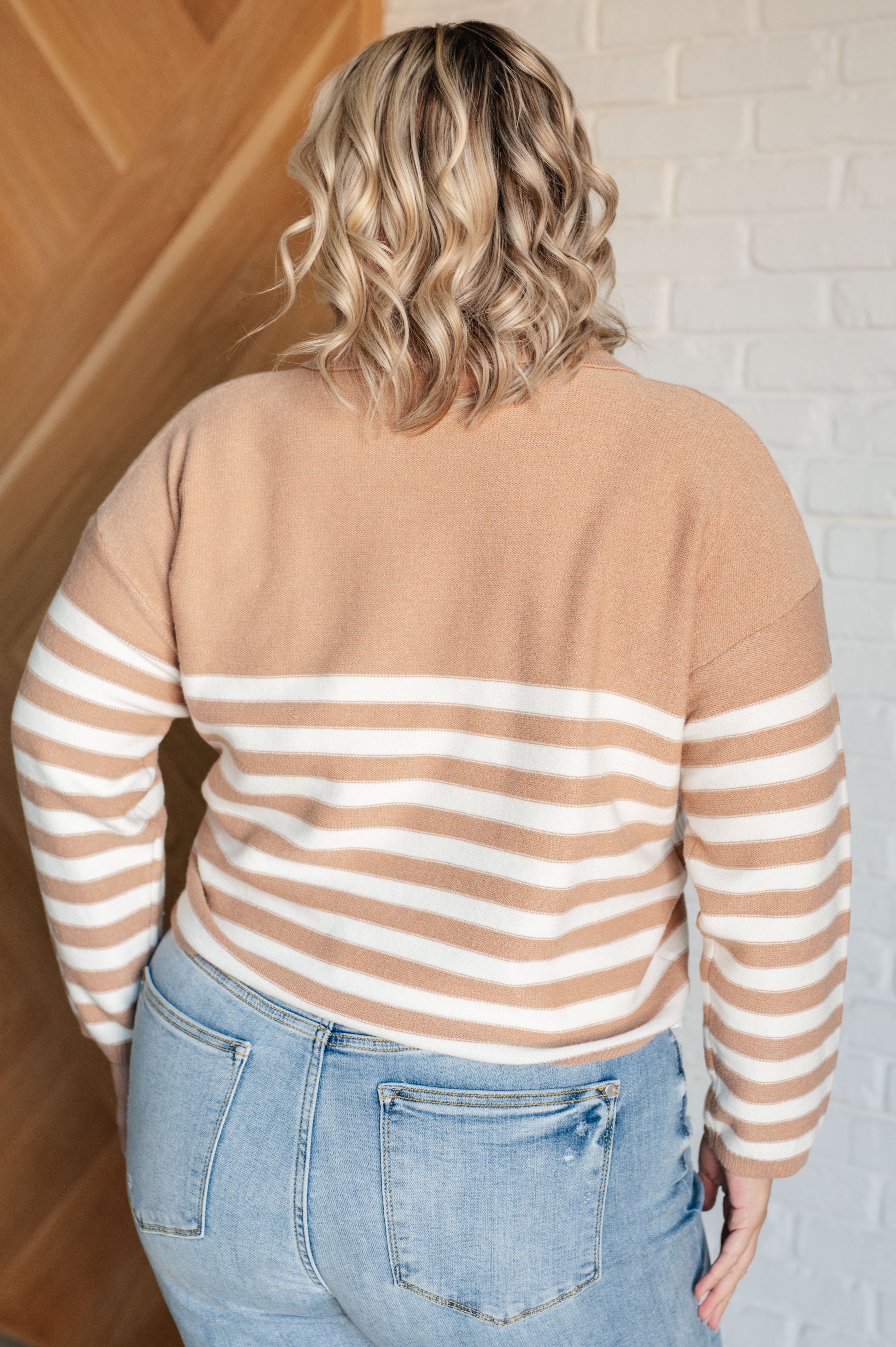 Person with wavy blonde hair wearing a mocha and white striped sweater with a relaxed fit, paired with light wash jeans, facing away indoors.