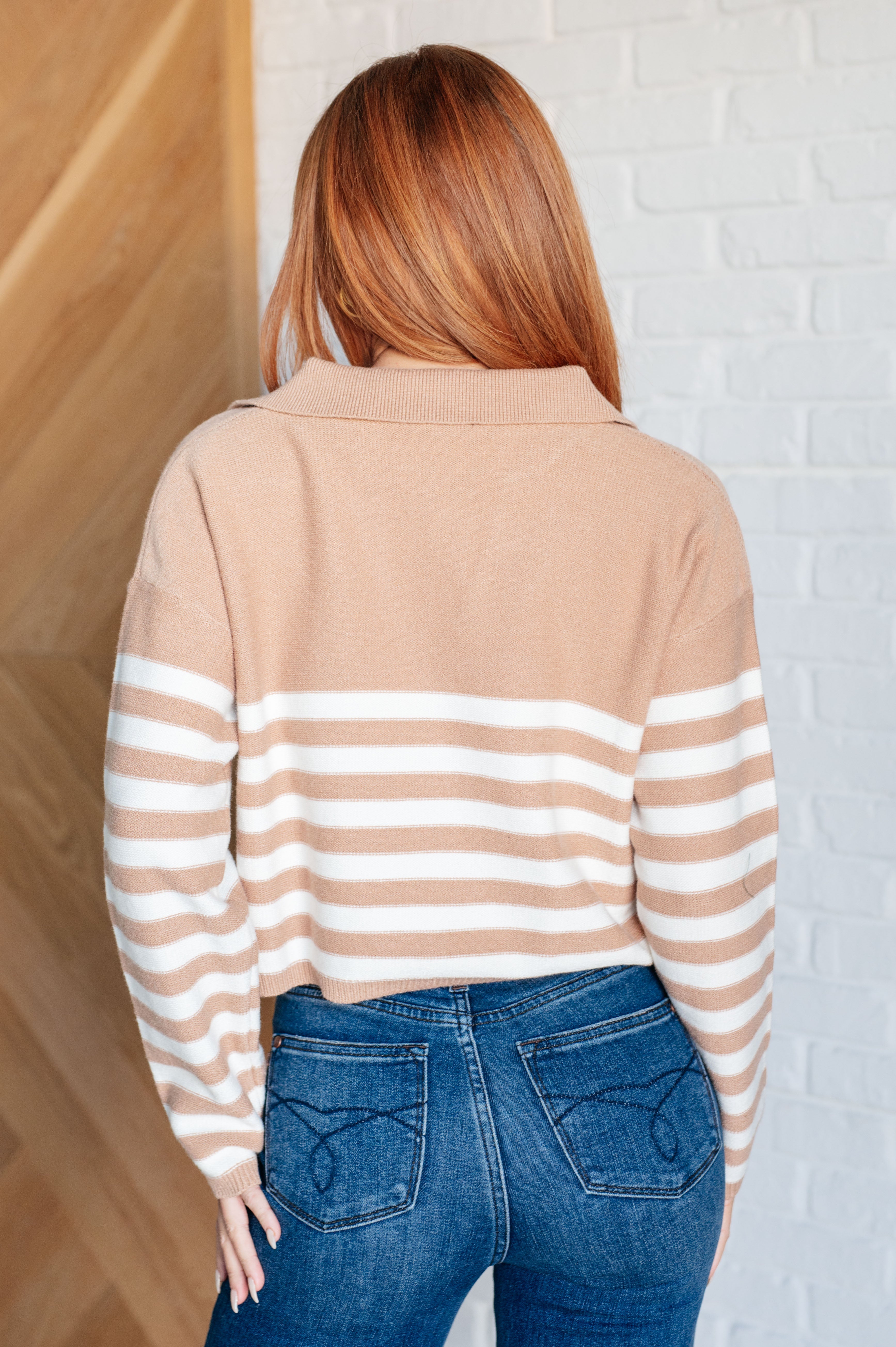 Woman with long red hair wearing a mocha striped sweater with horizontal white stripes on the sleeves and back, paired with dark blue jeans, standing indoors.