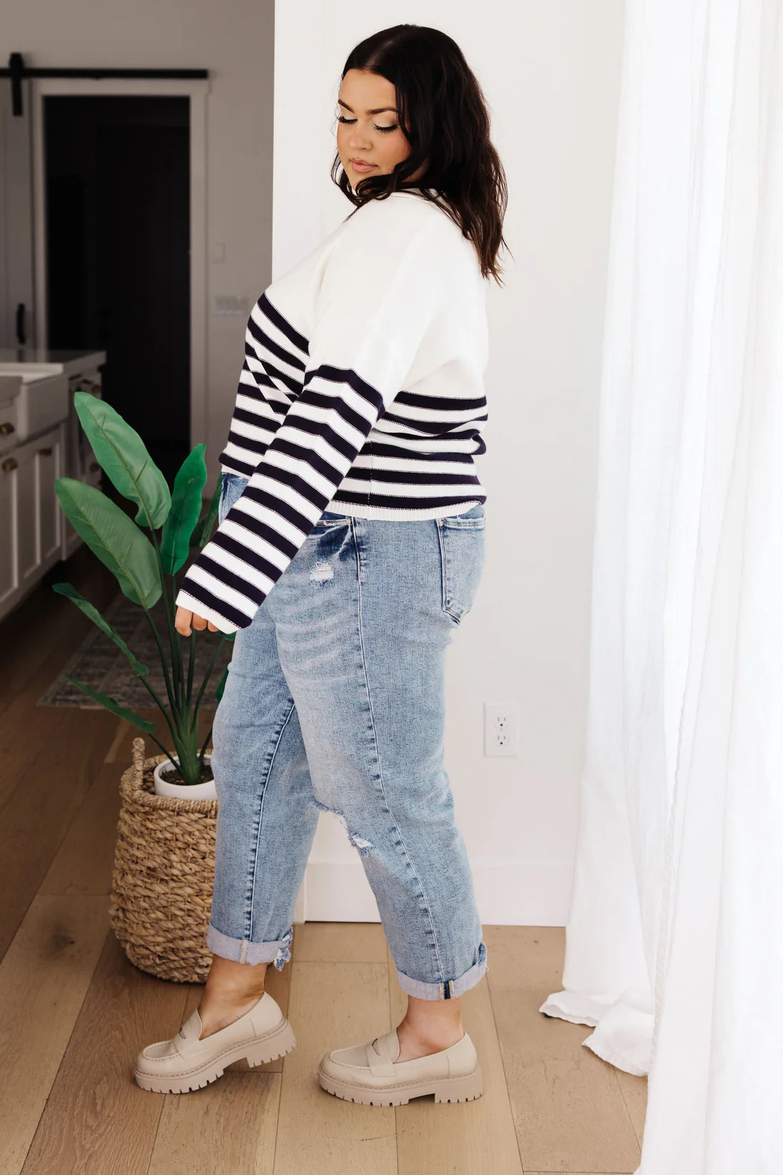 Woman with dark hair wearing a white sweater with black horizontal stripes on the lower half and sleeves, paired with cuffed light wash jeans and beige loafers, standing indoors near a plant.