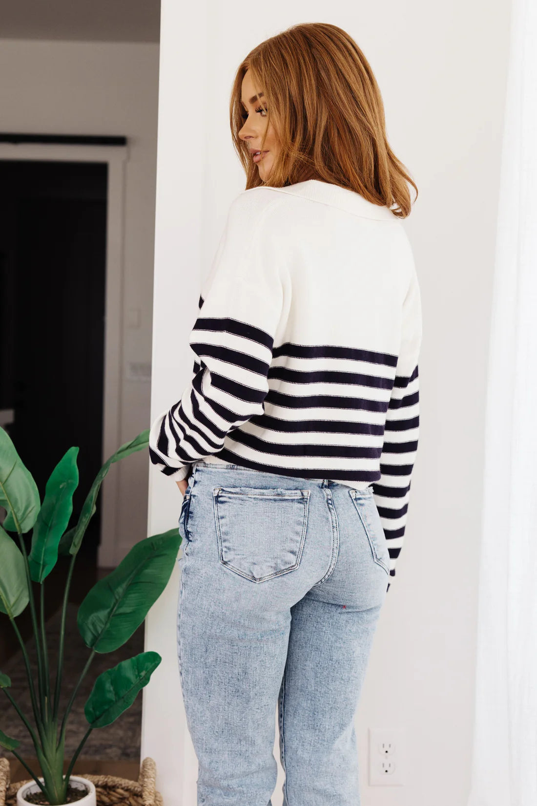 Woman with shoulder-length red hair wearing a white striped sweater with black horizontal stripes, paired with light wash high-rise jeans, standing indoors near a green plant.