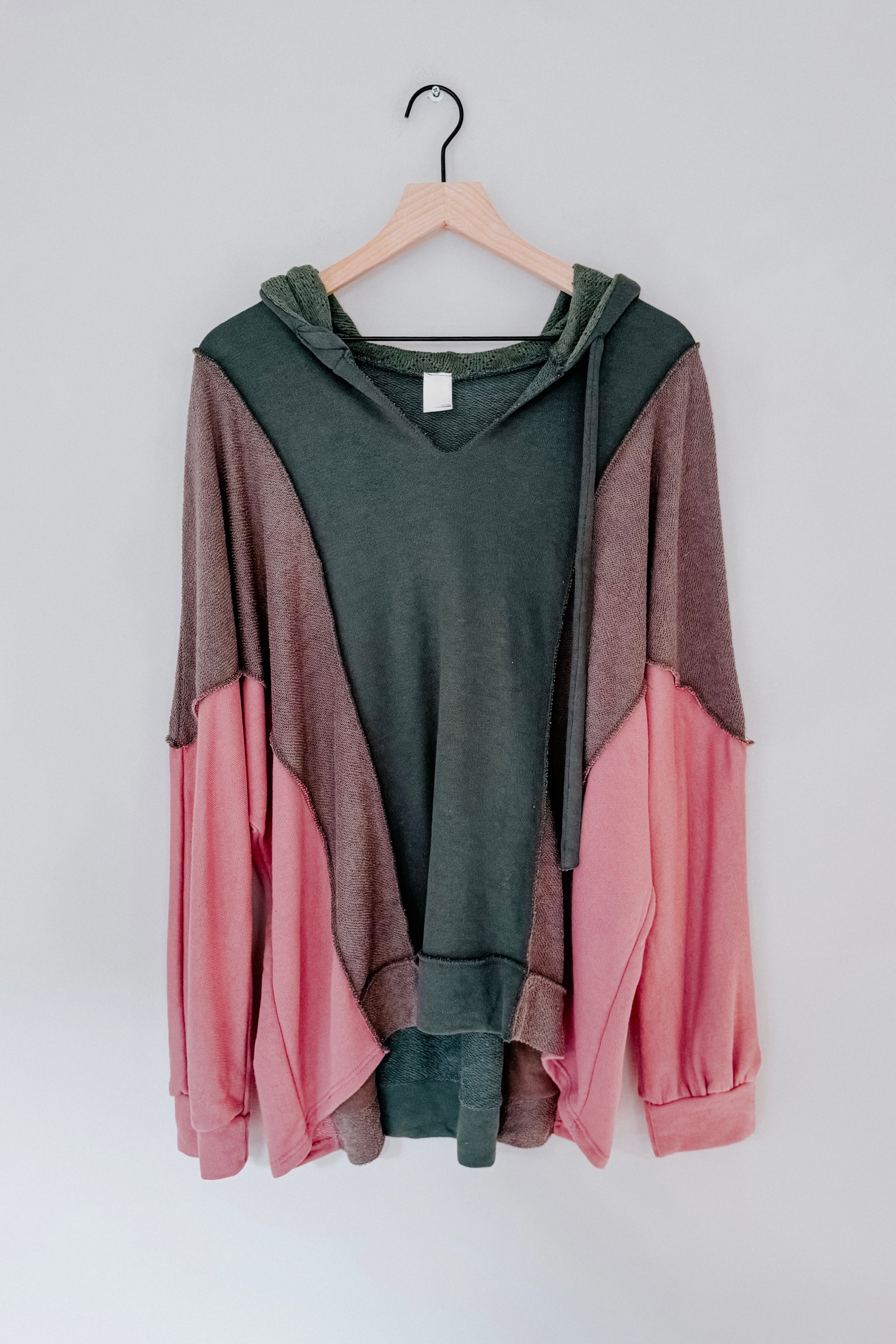Color block hoodie in sage green, muted brown, and soft pink hanging on a wooden hanger against a plain light background.