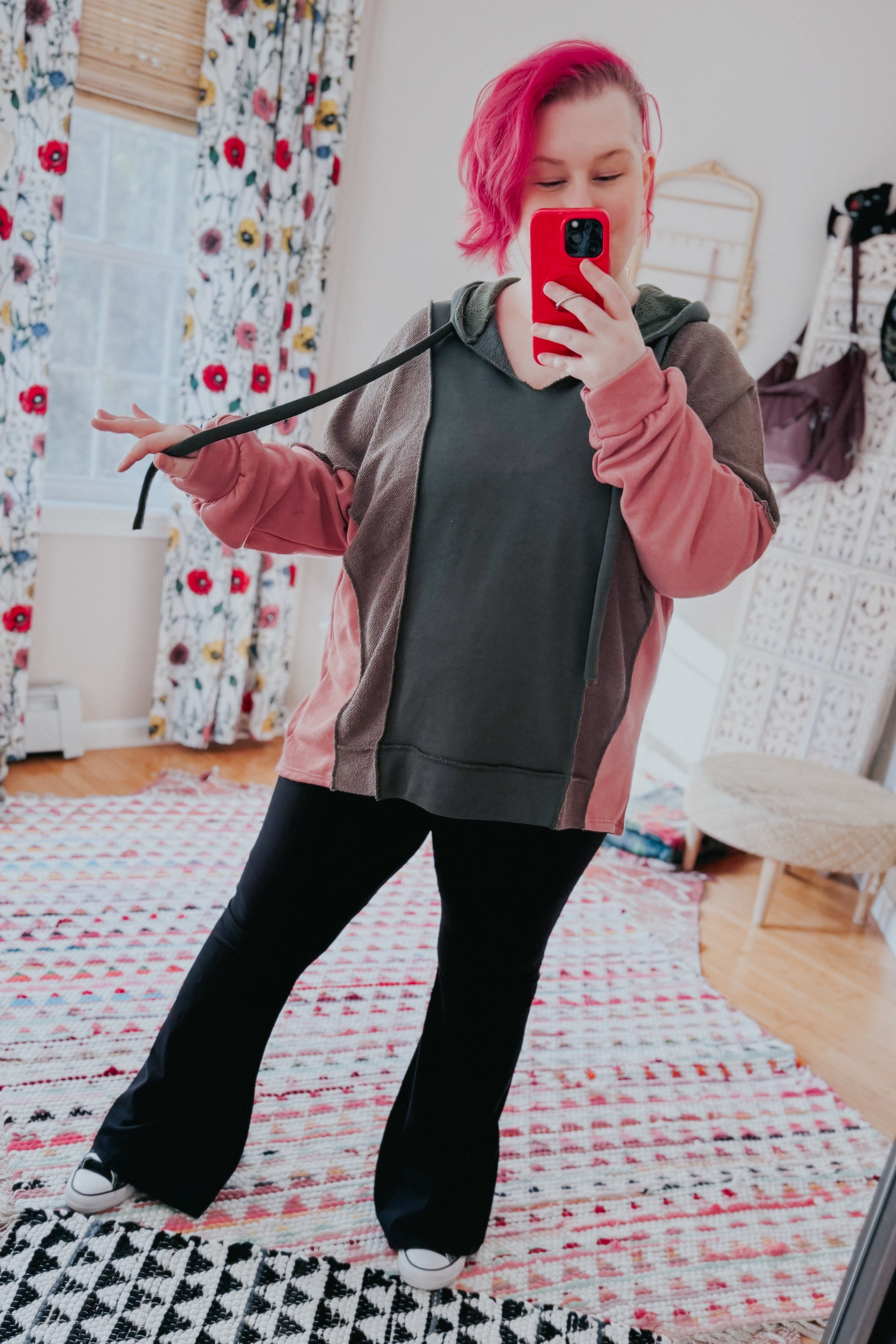 Woman with bright pink hair wearing a relaxed color block hoodie in sage, muted brown, and soft pink, taking a mirror selfie indoors on patterned rugs.