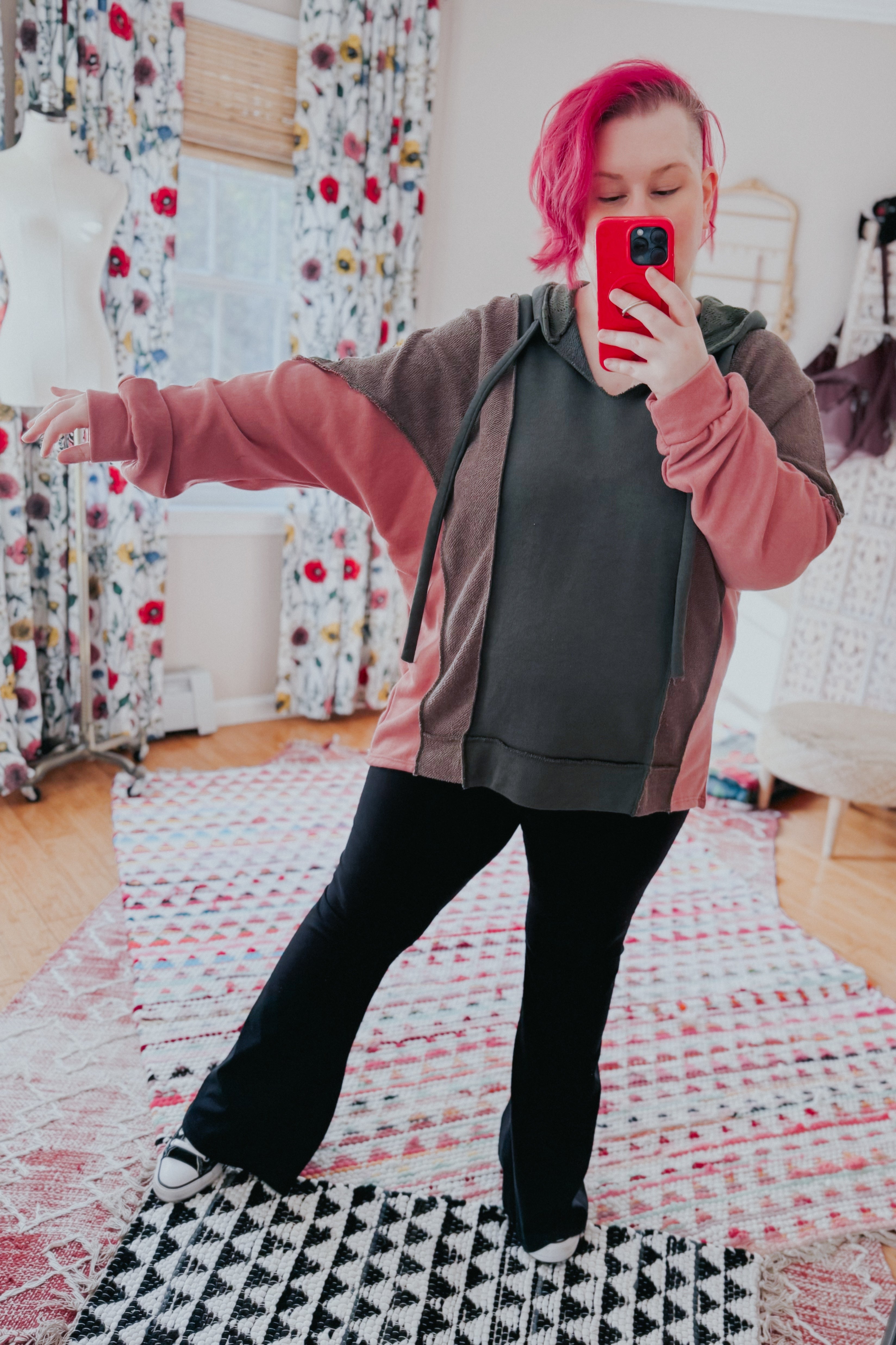 Woman with bright pink hair wearing a relaxed fit Message Me Back hoodie in sage, muted brown, and pink tones, taking a selfie indoors on patterned rugs.