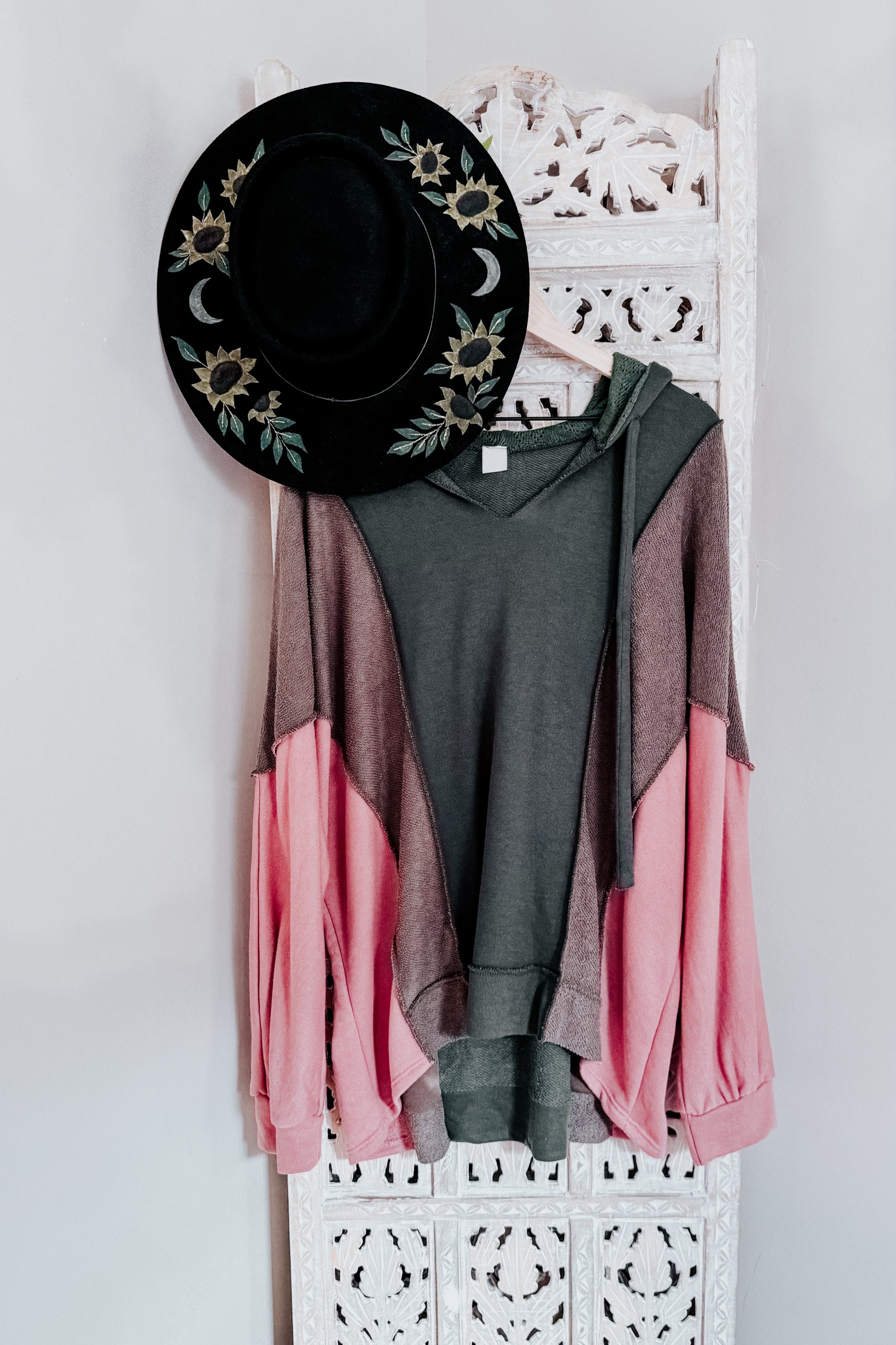 Sage hooded sweatshirt with muted brown and soft pink color blocks hanging on a carved white wooden panel beside a black hat with sunflower and moon designs.
