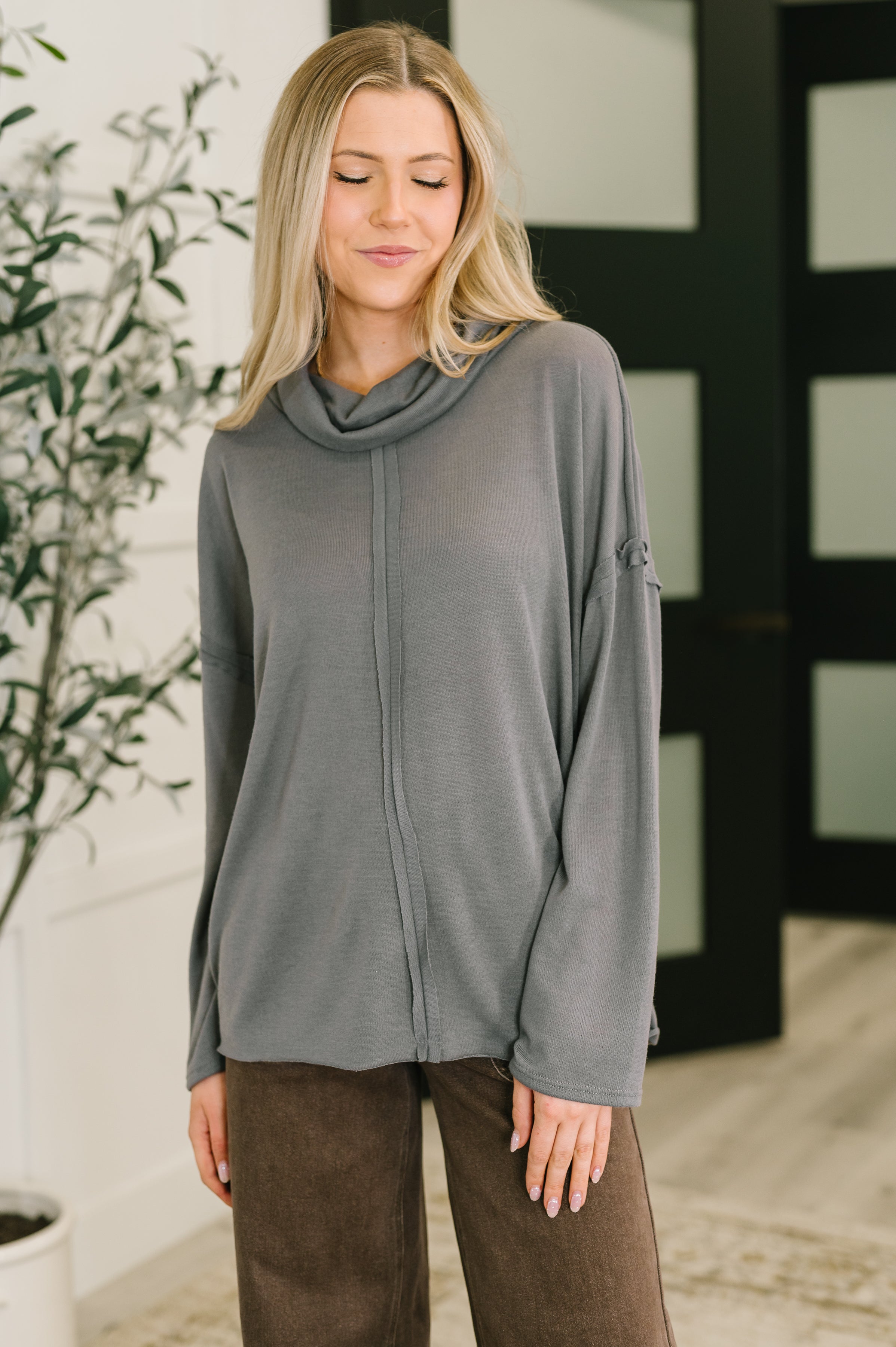Woman wearing a relaxed grey turtleneck top with a center seam detail, paired with brown pants in a softly lit modern room with plants.
