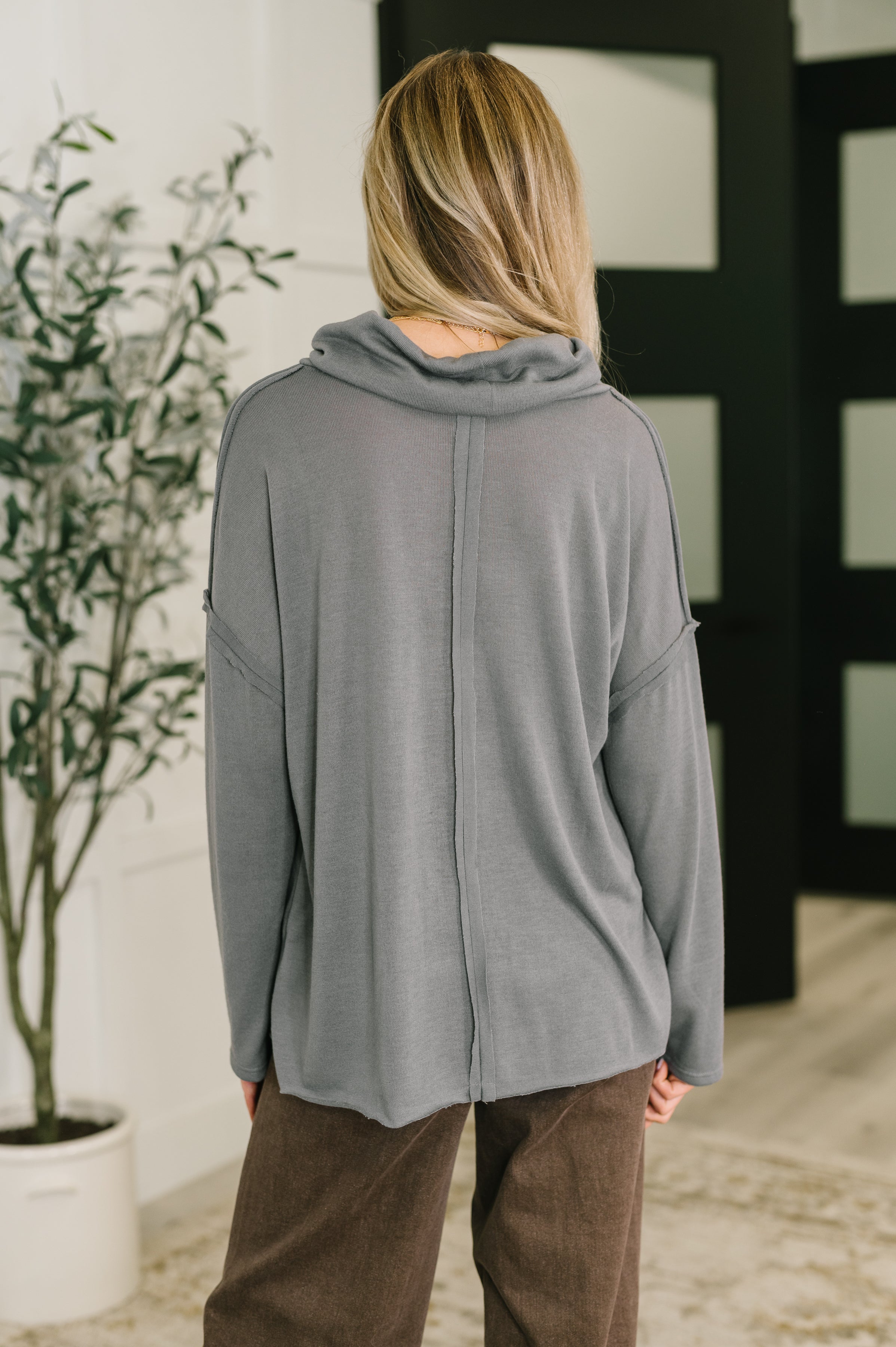 Rear view of a person wearing a loose-fitting grey turtleneck with a prominent center seam detail, paired with brown pants indoors.