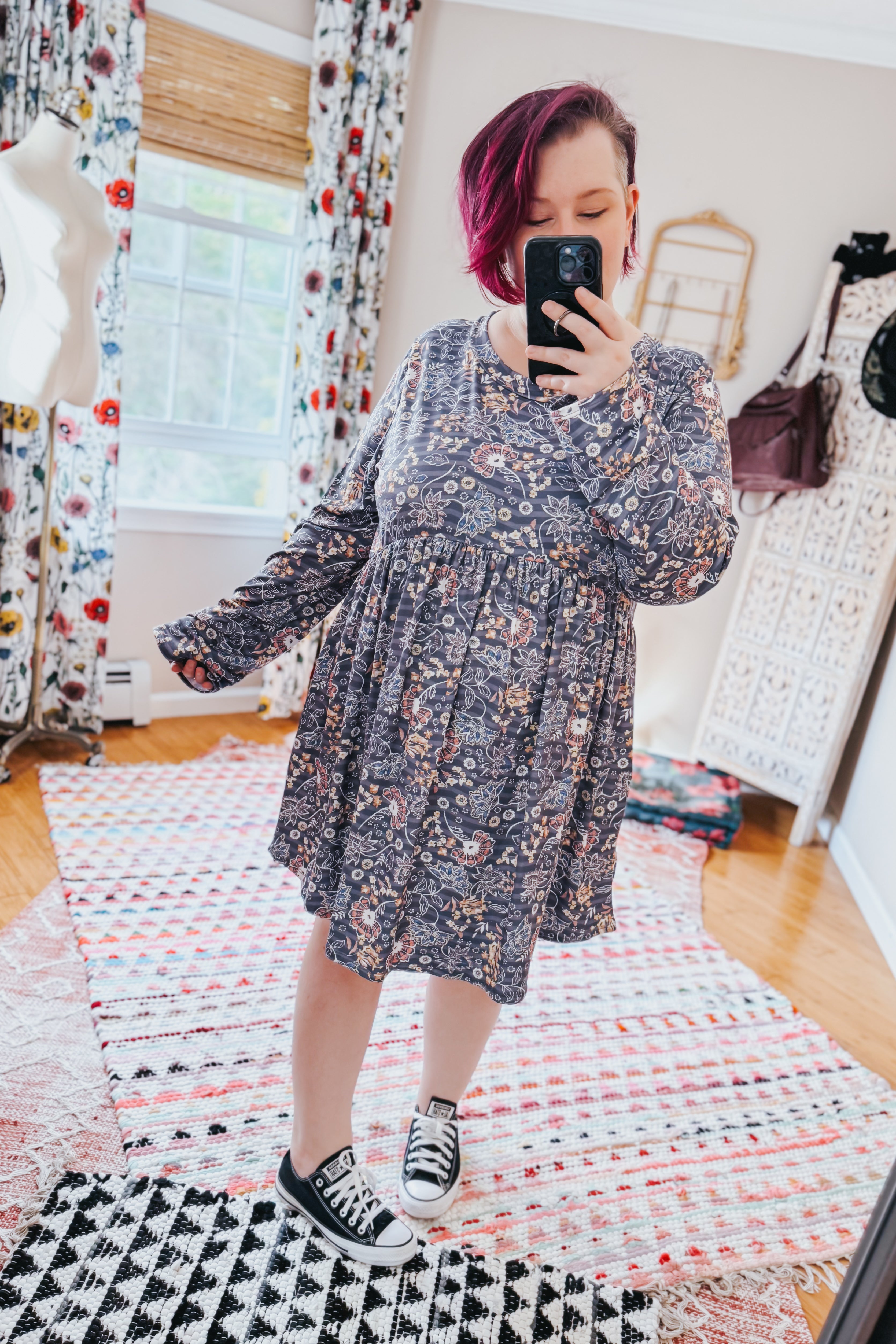 Woman taking a mirror selfie wearing a loose-fitting blue floral patterned swing dress with long sleeves in a bright room with patterned rugs and curtains.