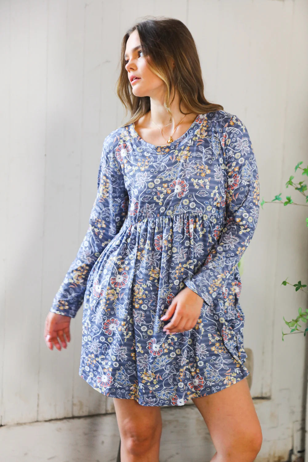 Young woman wearing a blue floral patterned swing dress with long sleeves and a gathered waist standing indoors near a light-colored wall and greenery.