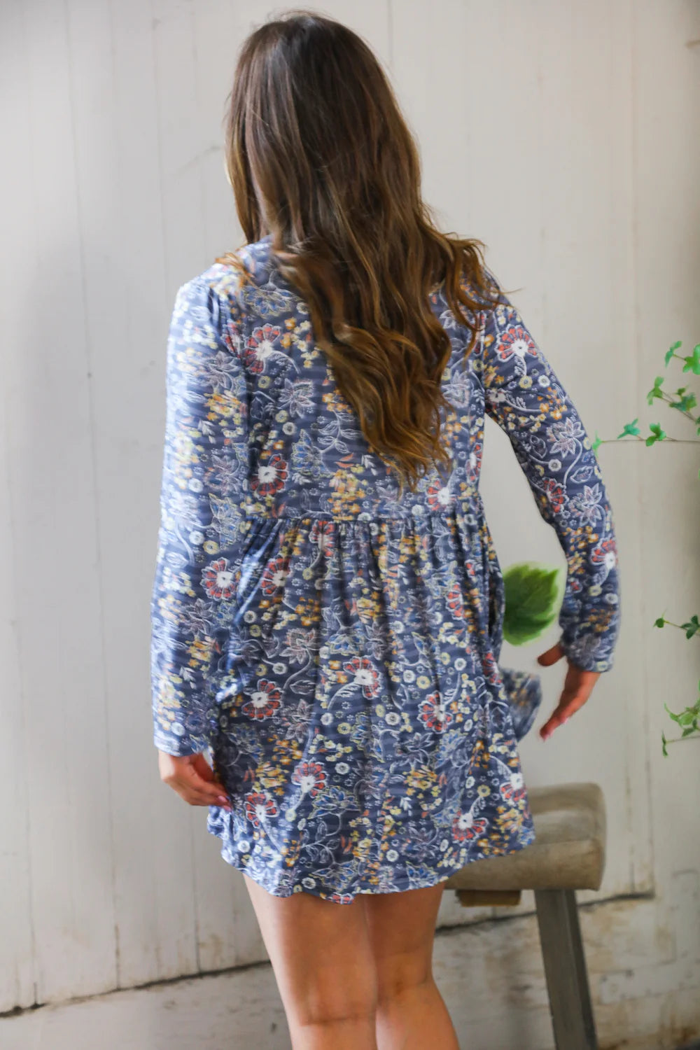 Woman seen from behind wearing a blue floral patterned swing dress with long sleeves, standing indoors near a wooden stool and light-colored wall.