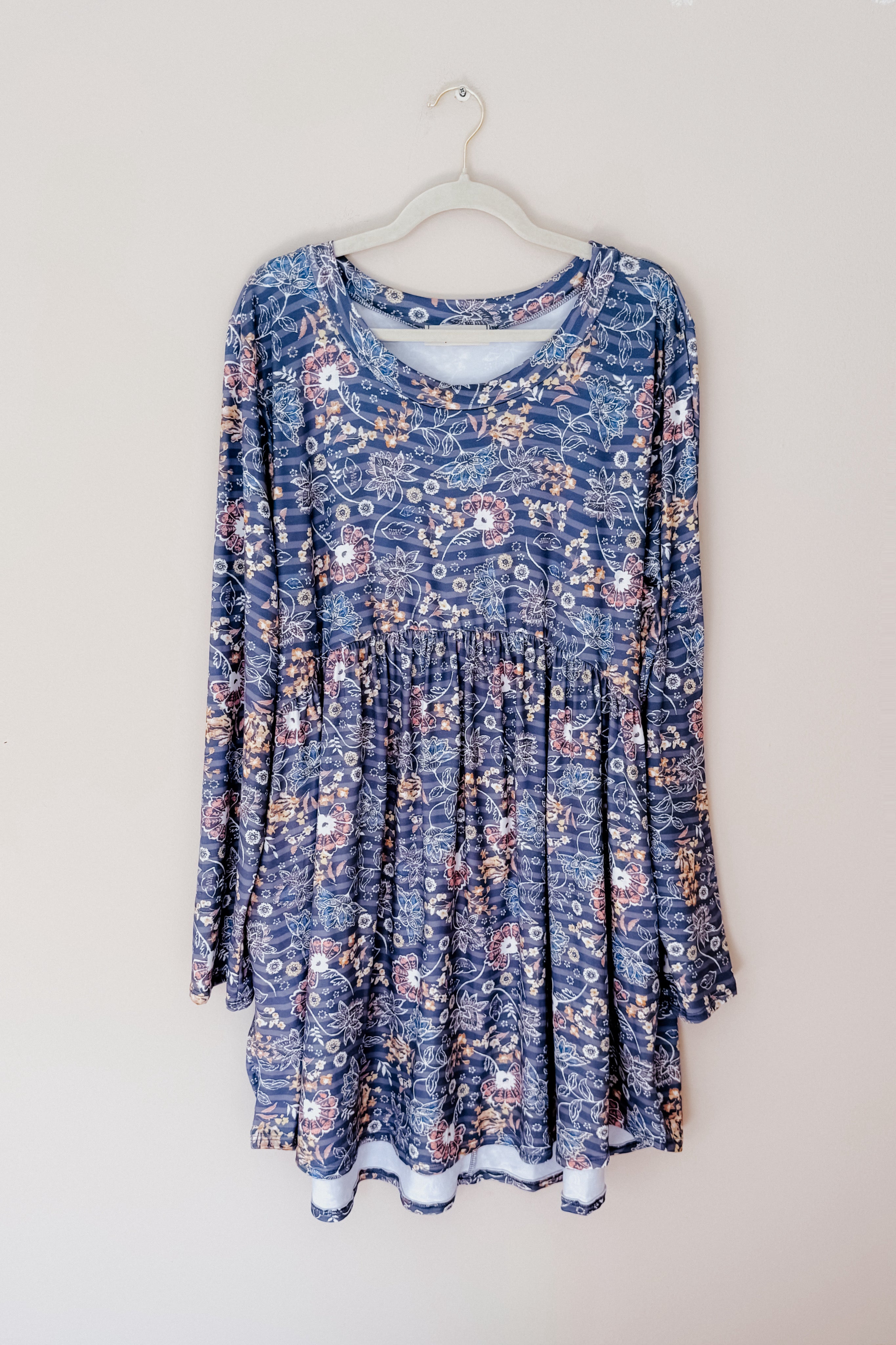 Blue long-sleeve dress with a floral pattern and gathered waist hanging on a white hanger against a light wall background